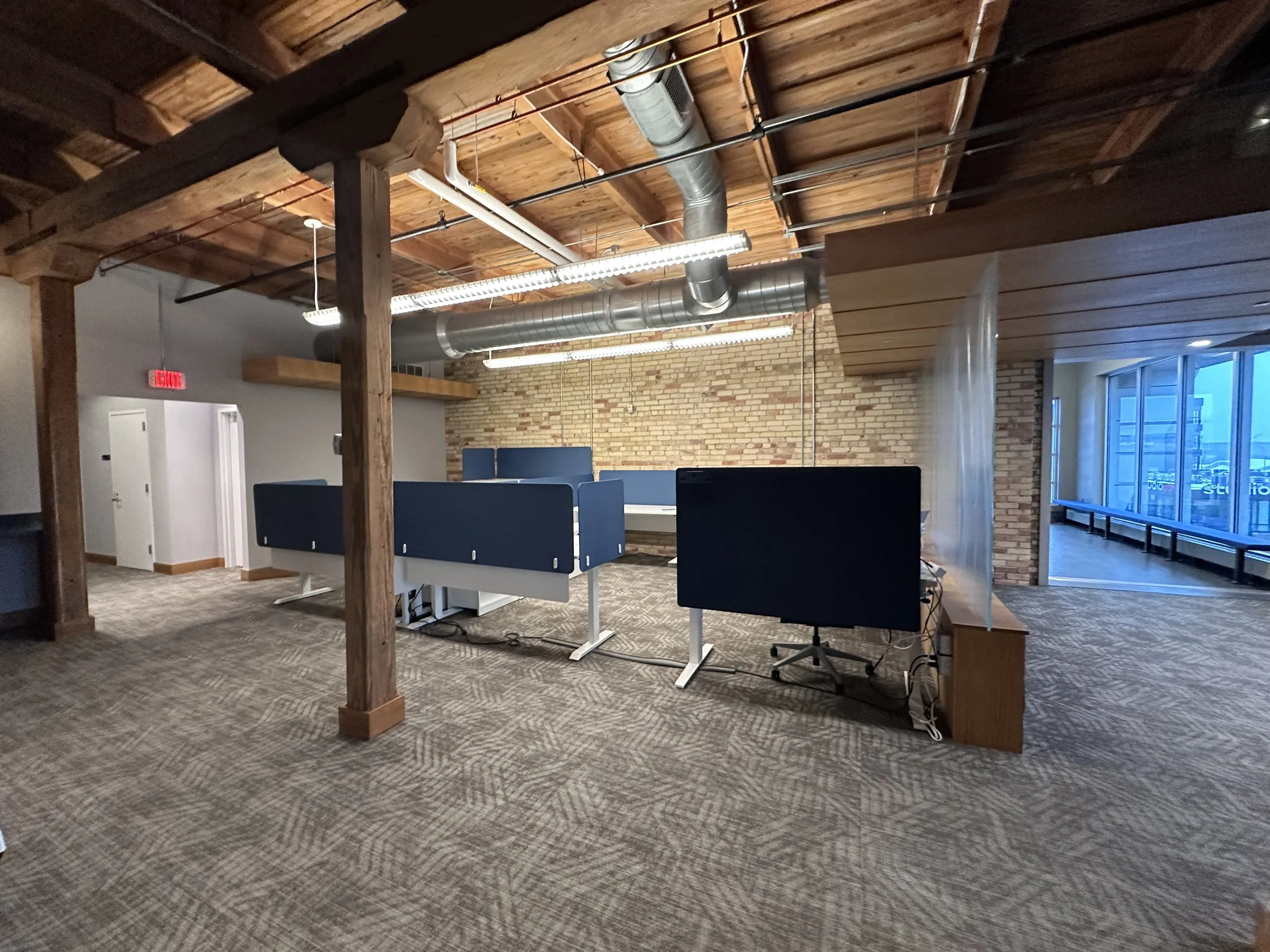 Office with exposed brick wall, wooden ceiling beams, ductwork, workstations with black monitors, and large windows.