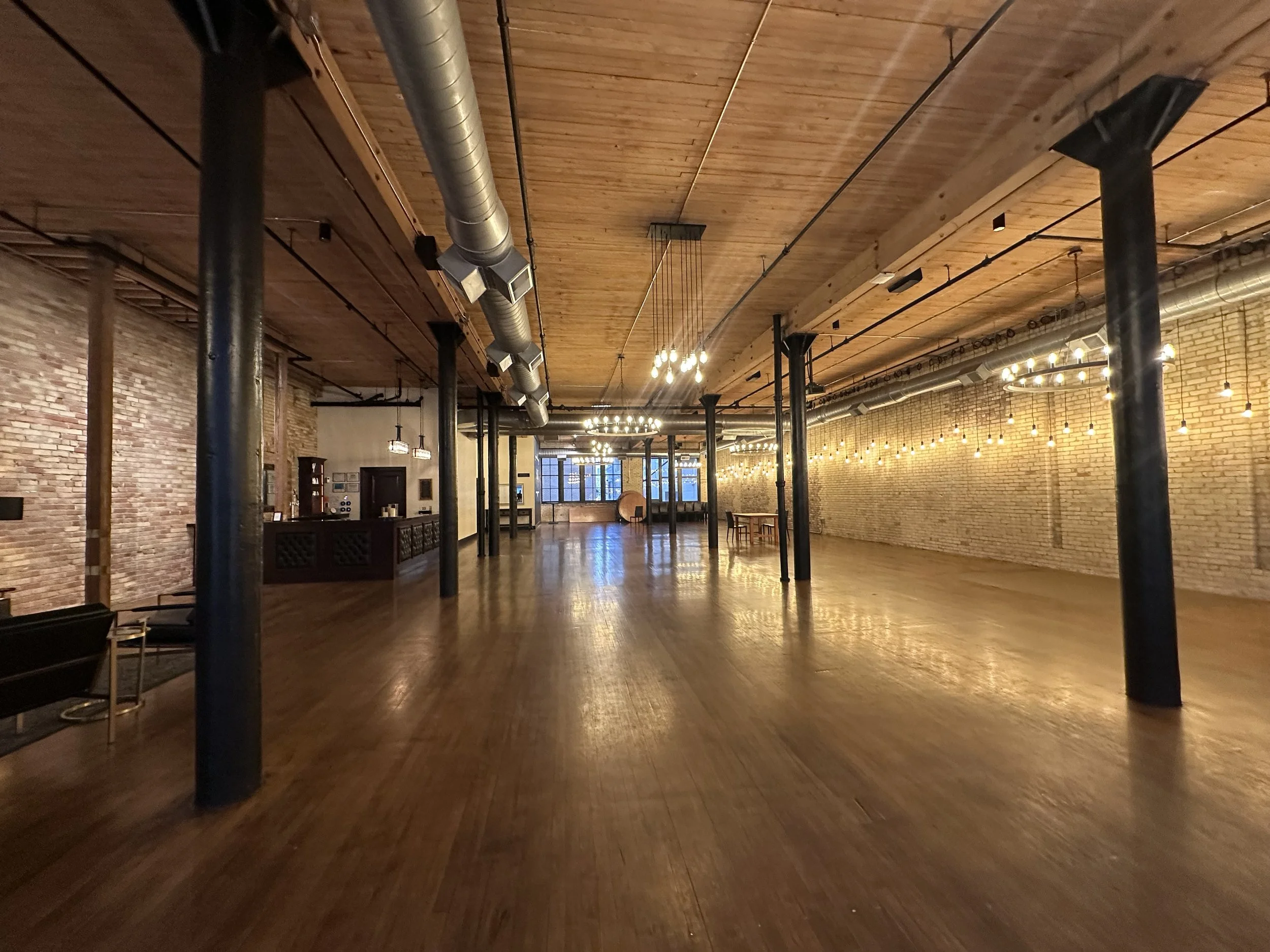 Spacious empty event or dance hall with wooden floors, brick walls, and string lights hanging from the ceiling. There are black metal support beams, industrial ductwork, and a reception counter on the left side.