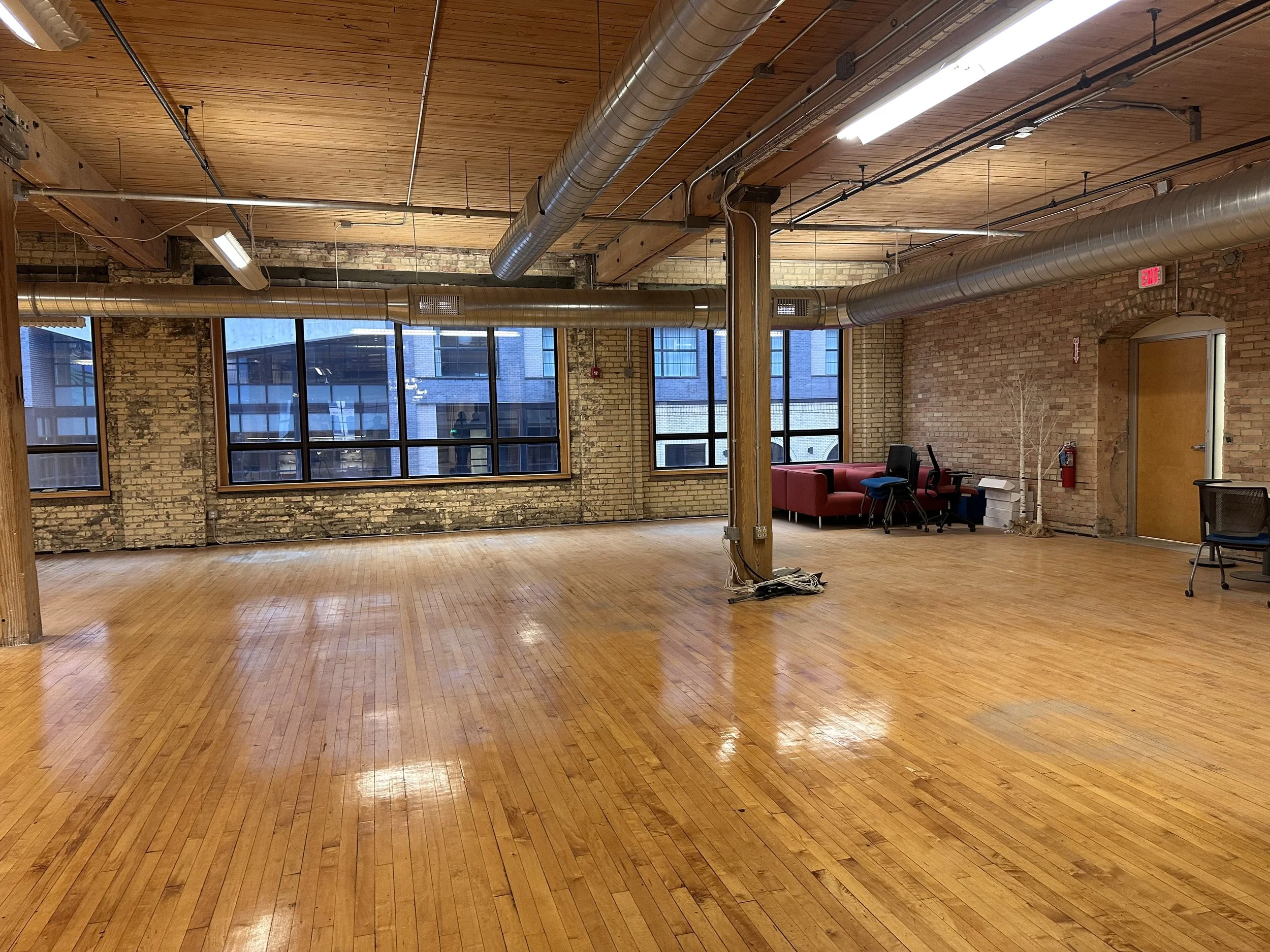 Spacious indoor room with wooden floors and brick walls, large windows, exposed ductwork, and minimal furniture including chairs, a sofa, and a fire extinguisher.