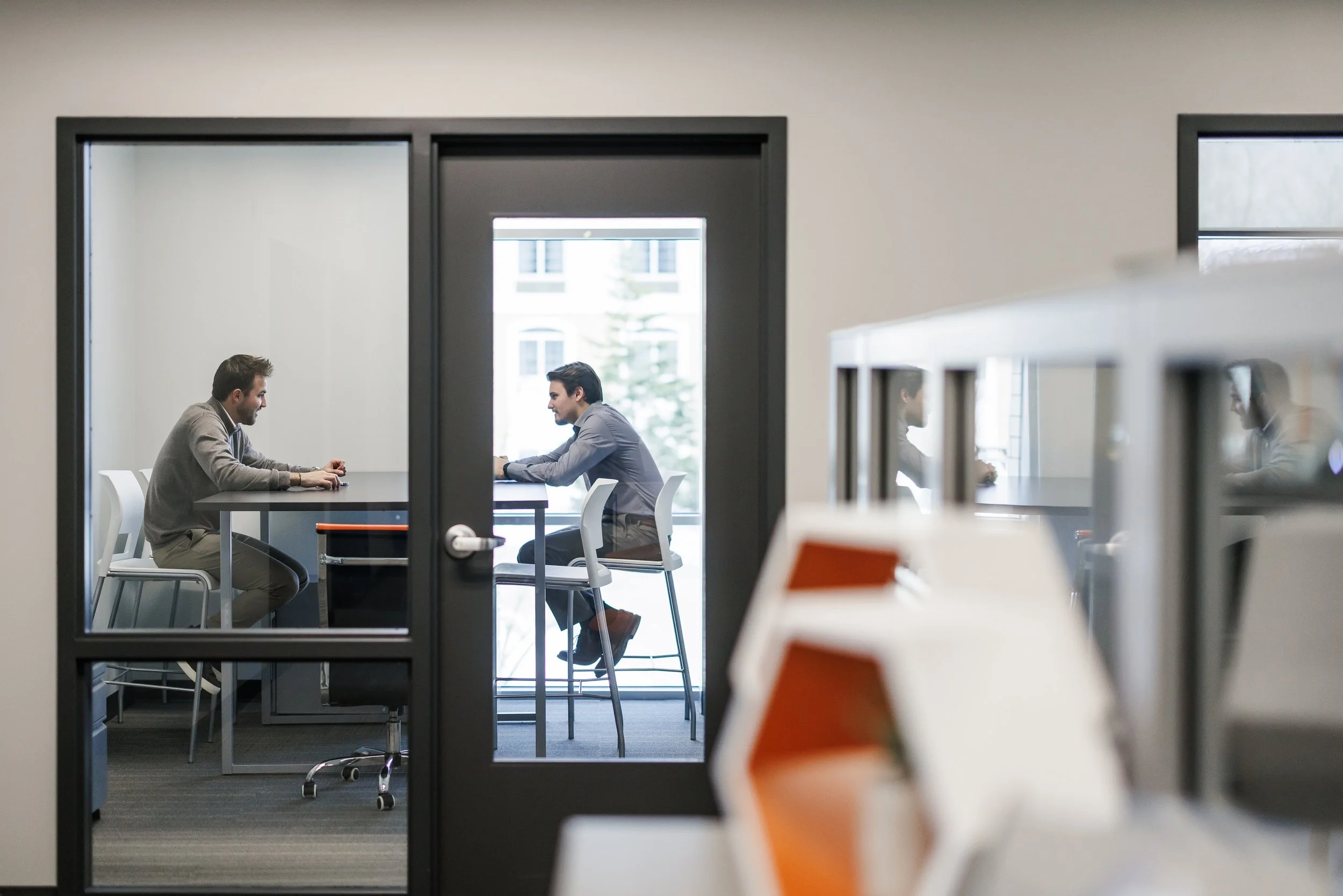 Two men having a meeting in a modern office conference room, seen through a glass door, with a view of desks and office furniture in the foreground.