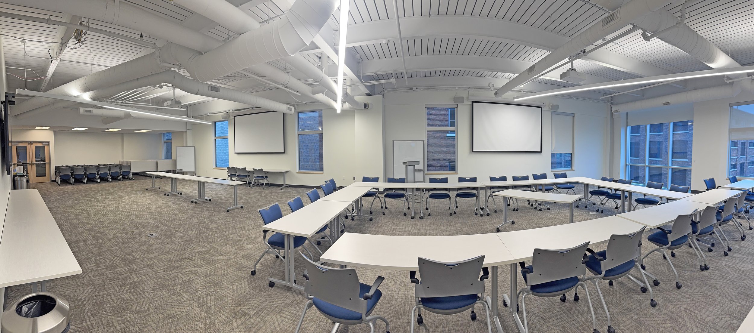 A modern, spacious conference room with white walls, multiple windows, and a high ceiling with exposed ductwork. There are several white tables arranged in a U-shape with blue and white chairs around them. Two large projection screens are mounted on the front wall, with a podium and whiteboard nearby.