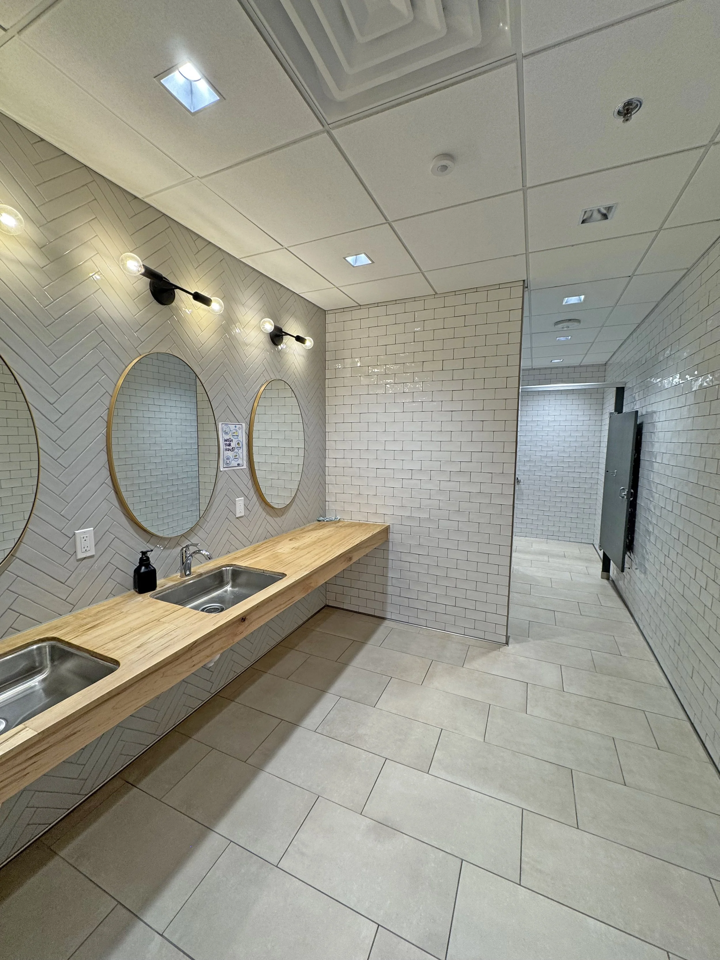 Public restroom with two sinks, wooden counter, oval mirrors, tiled wall, and a mirror partition. The ceiling has recessed lighting and a vent.