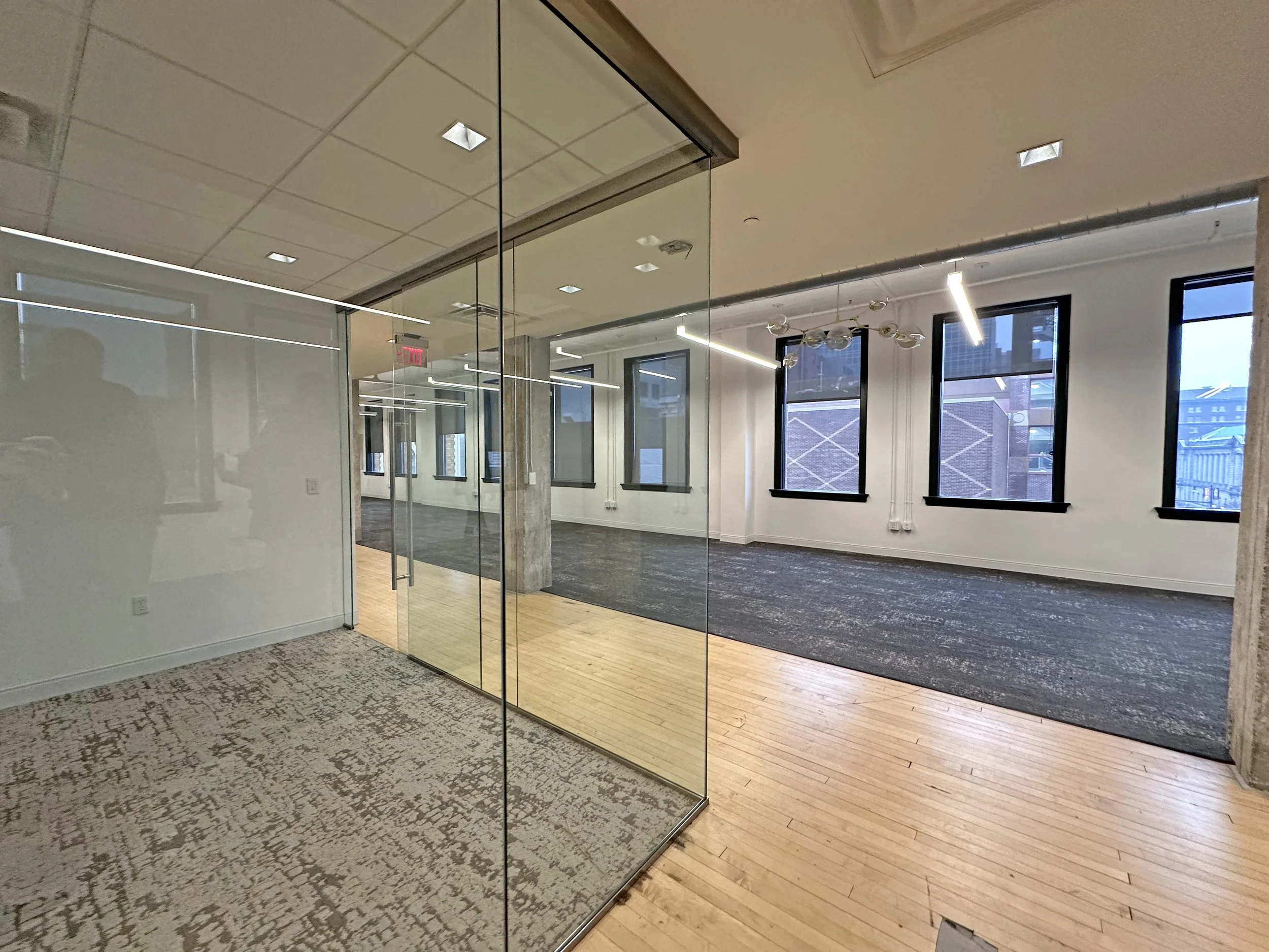 Empty modern office space with large windows, black window frames, and wooden flooring, divided by glass walls. The area features contemporary lighting fixtures and a patterned rug.