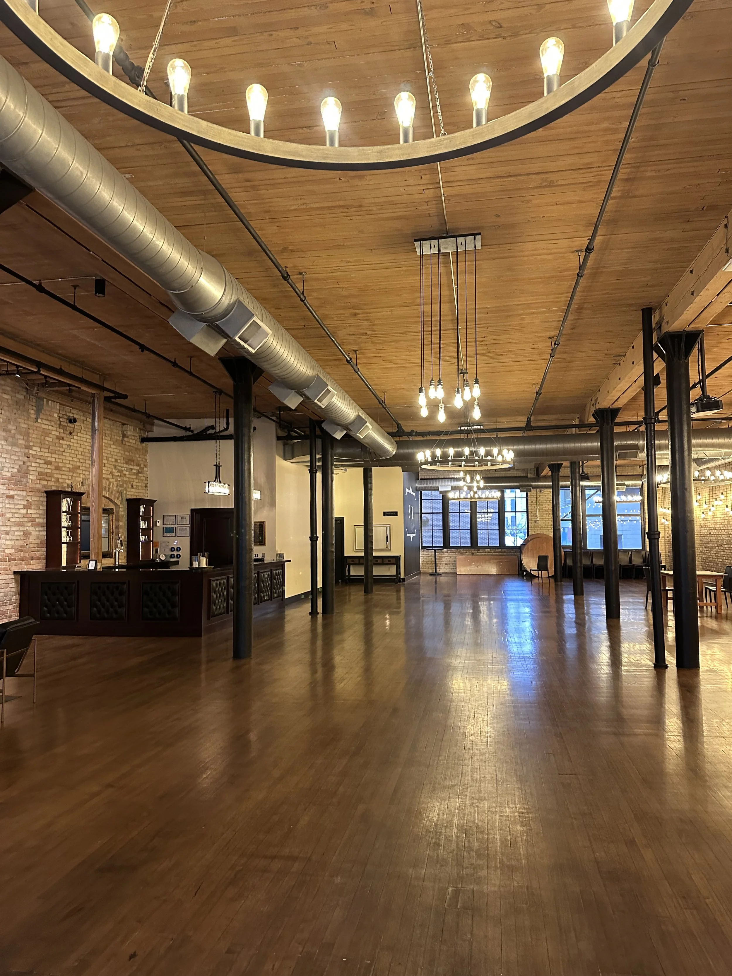 Empty event space with wooden floors, brick walls, industrial pipes, and warm lighting fixtures hanging from a wooden ceiling.