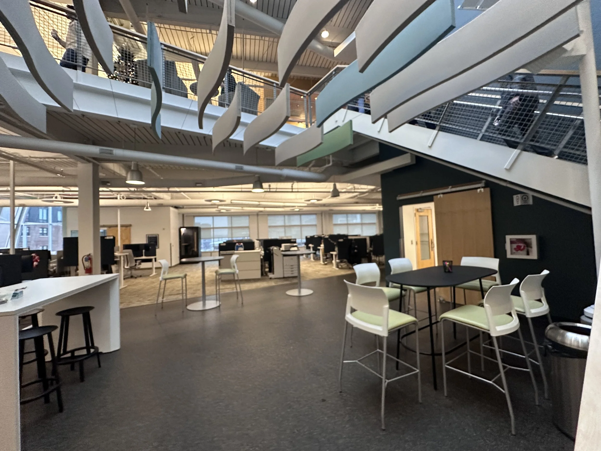 Open office space with desks, chairs, and computer monitors, with a mezzanine level above with people walking and talking.