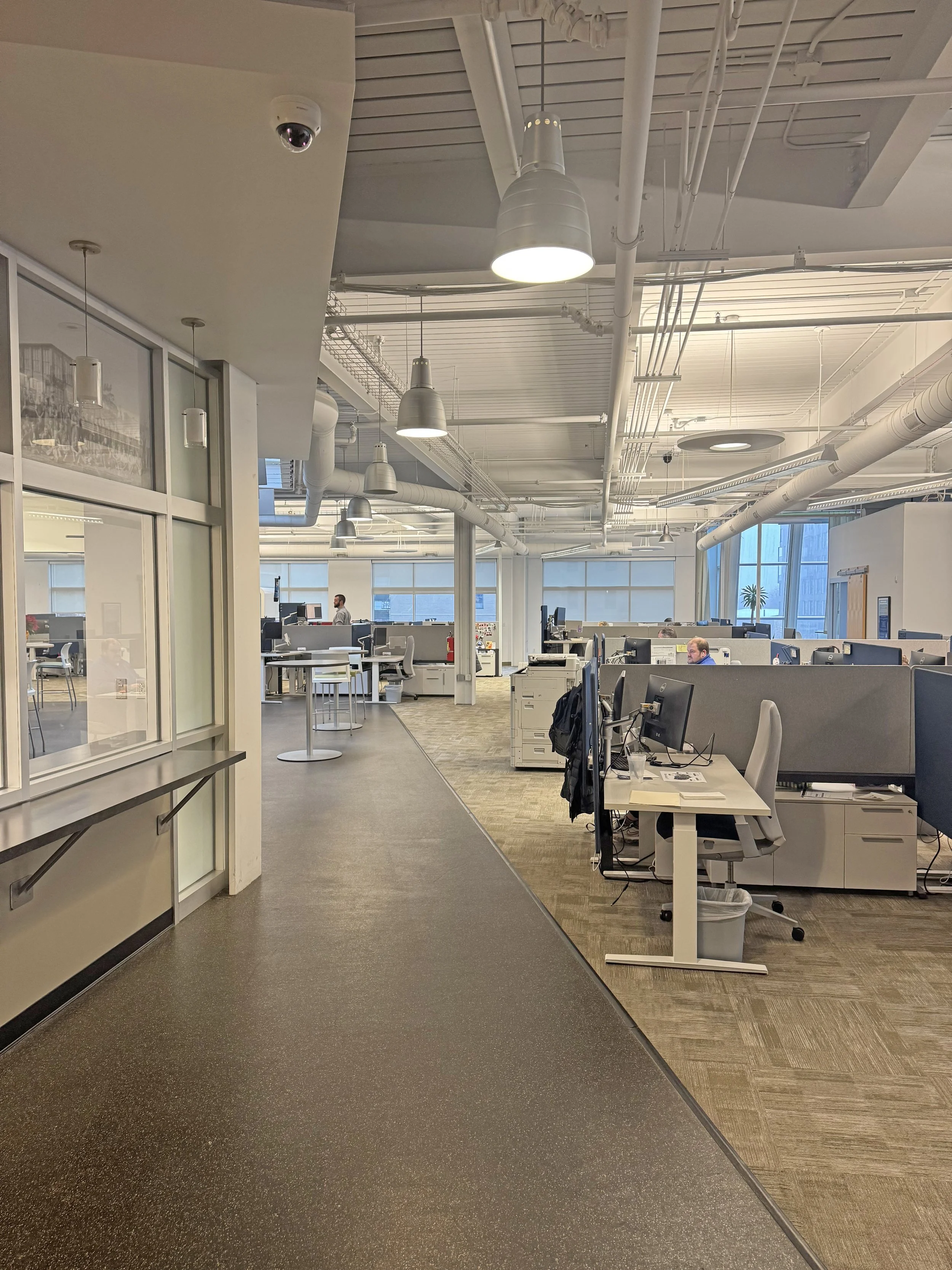 Open office space with cubicles, desks, and computers, large windows with blinds, and ceiling with exposed pipes and ducts.