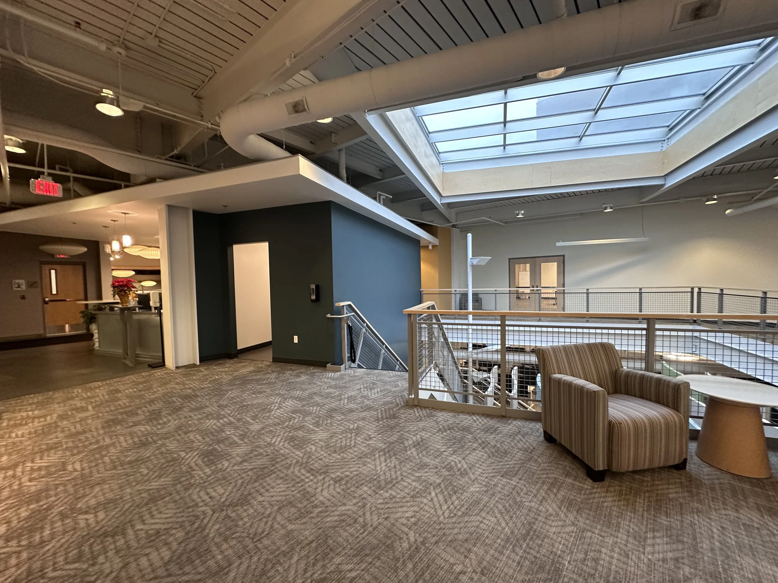 Interior of a multi-story building with an open atrium, seating area with an armchair and side table, skylight ceiling, staircase, and hallway leading to other rooms.