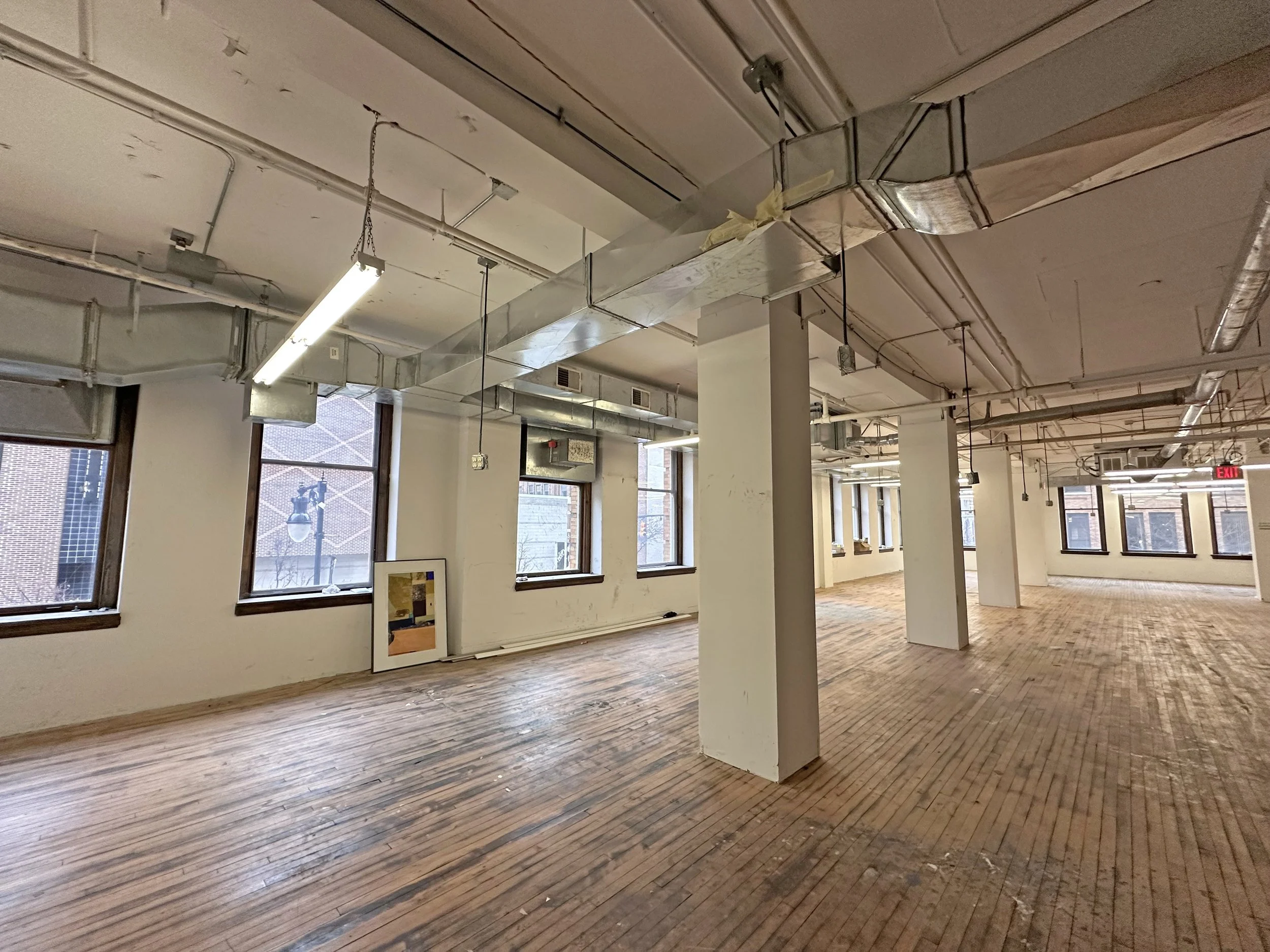 Empty commercial or office space with large windows, wooden floors, exposed ductwork, and white walls.