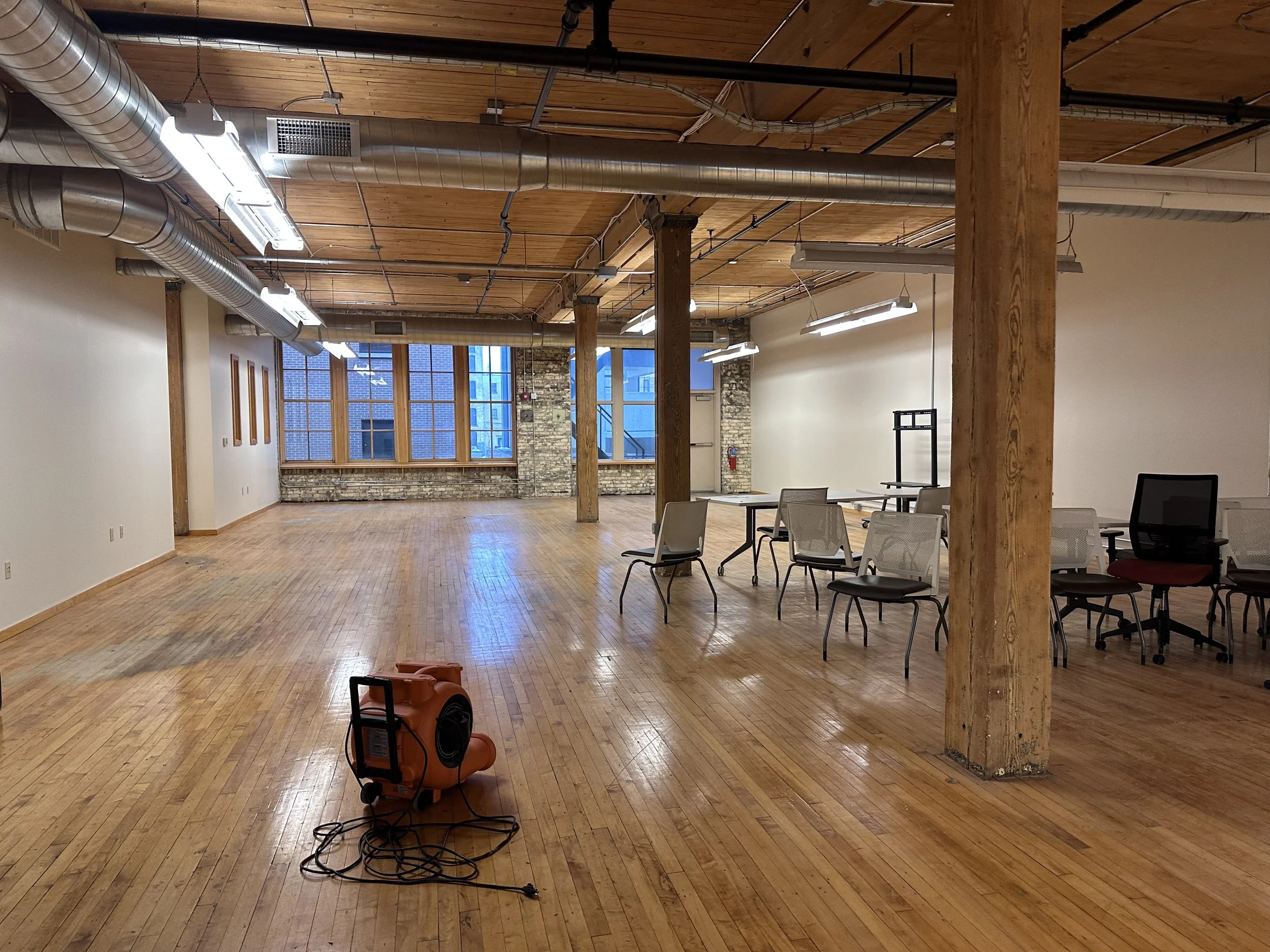 Empty room with wooden floors and exposed brick wall at the far end, large windows, several chairs, and a portable heater on the floor.