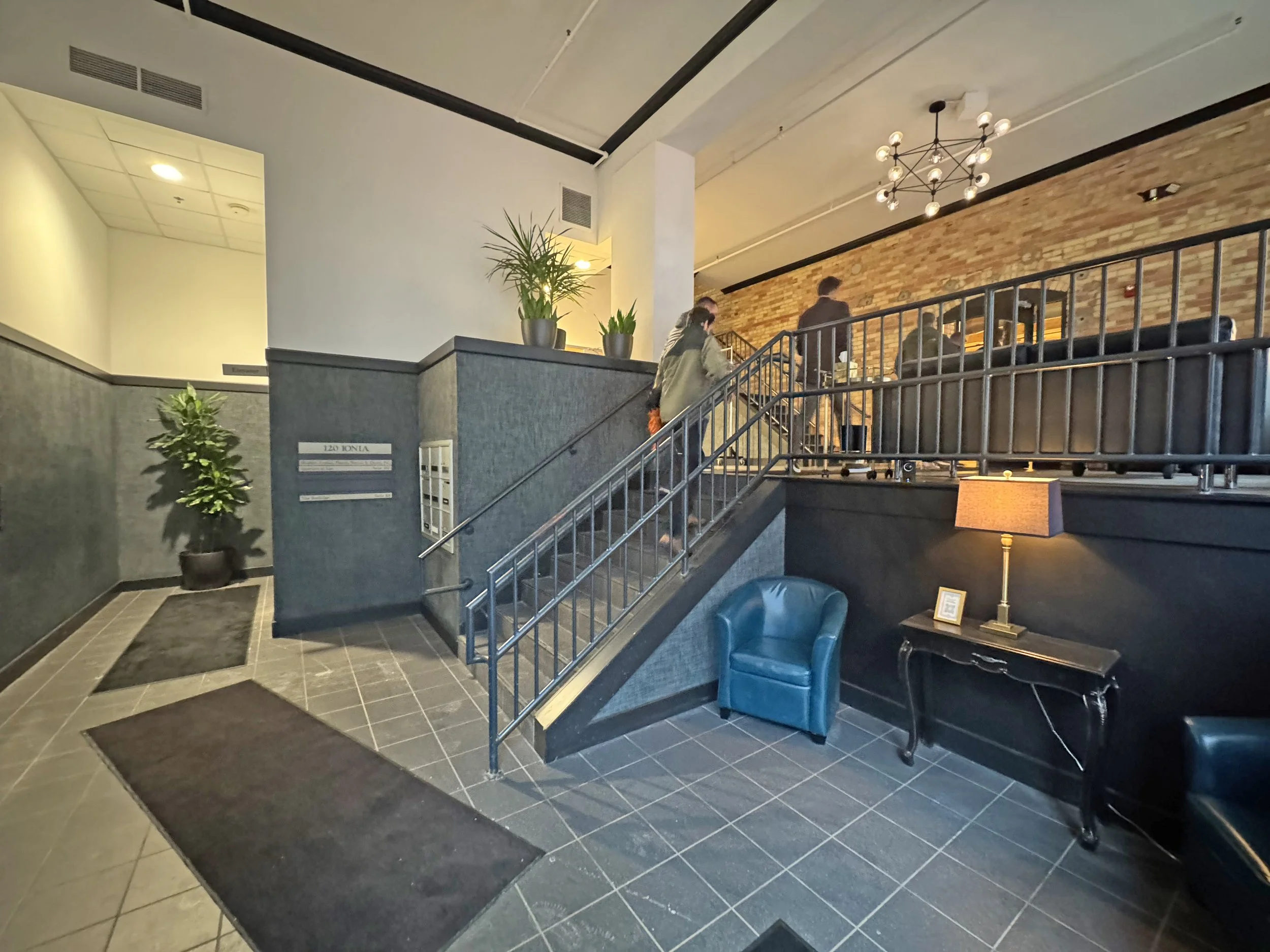 Interior of a modern building with a staircase, potted plants, a black table with a lamp, and people walking up the stairs.