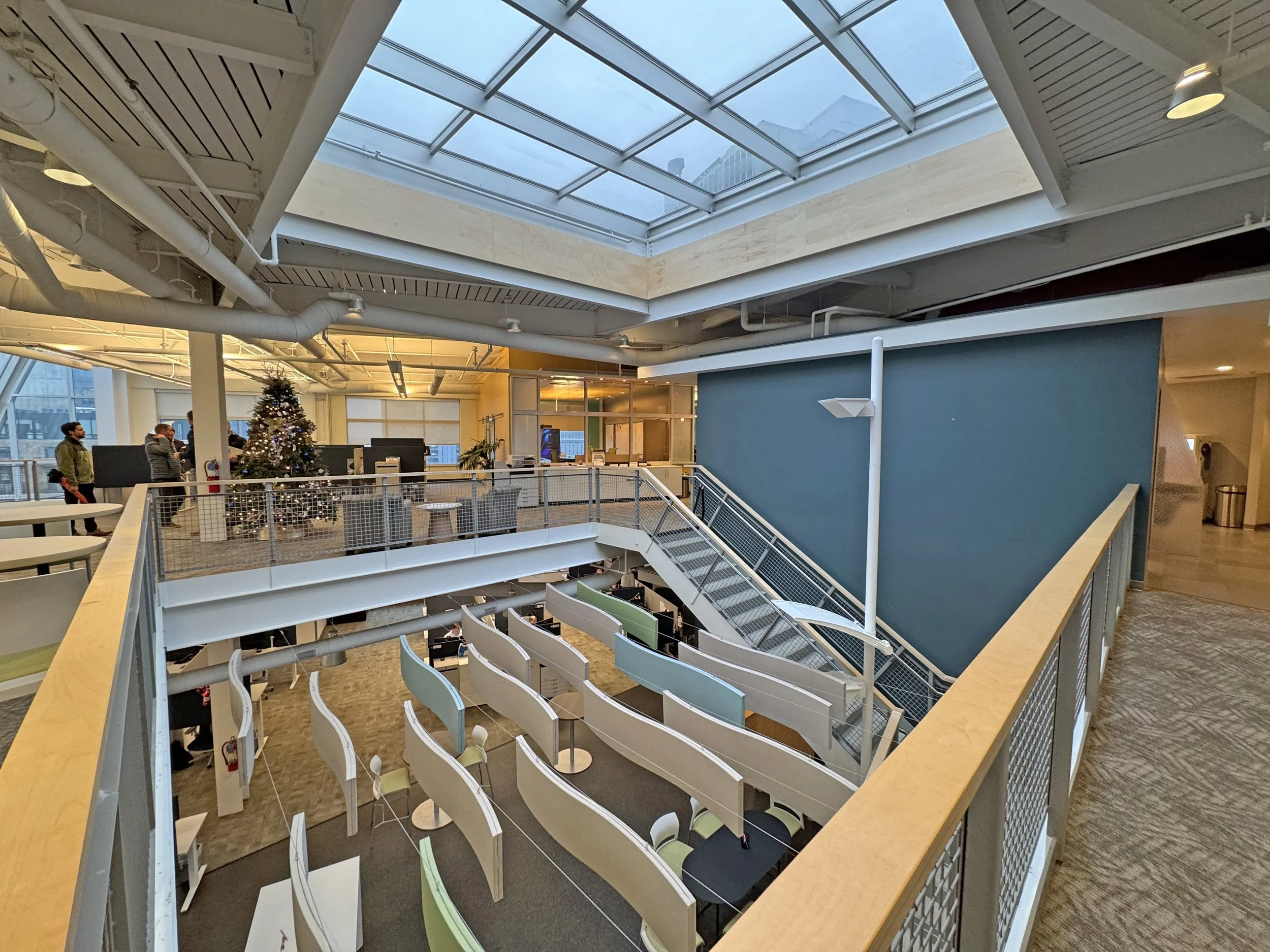 Interior of a modern office space with a large skylight, a decorated Christmas tree, a few people walking, and a staircase leading to a lower level with cubicles and workstations.