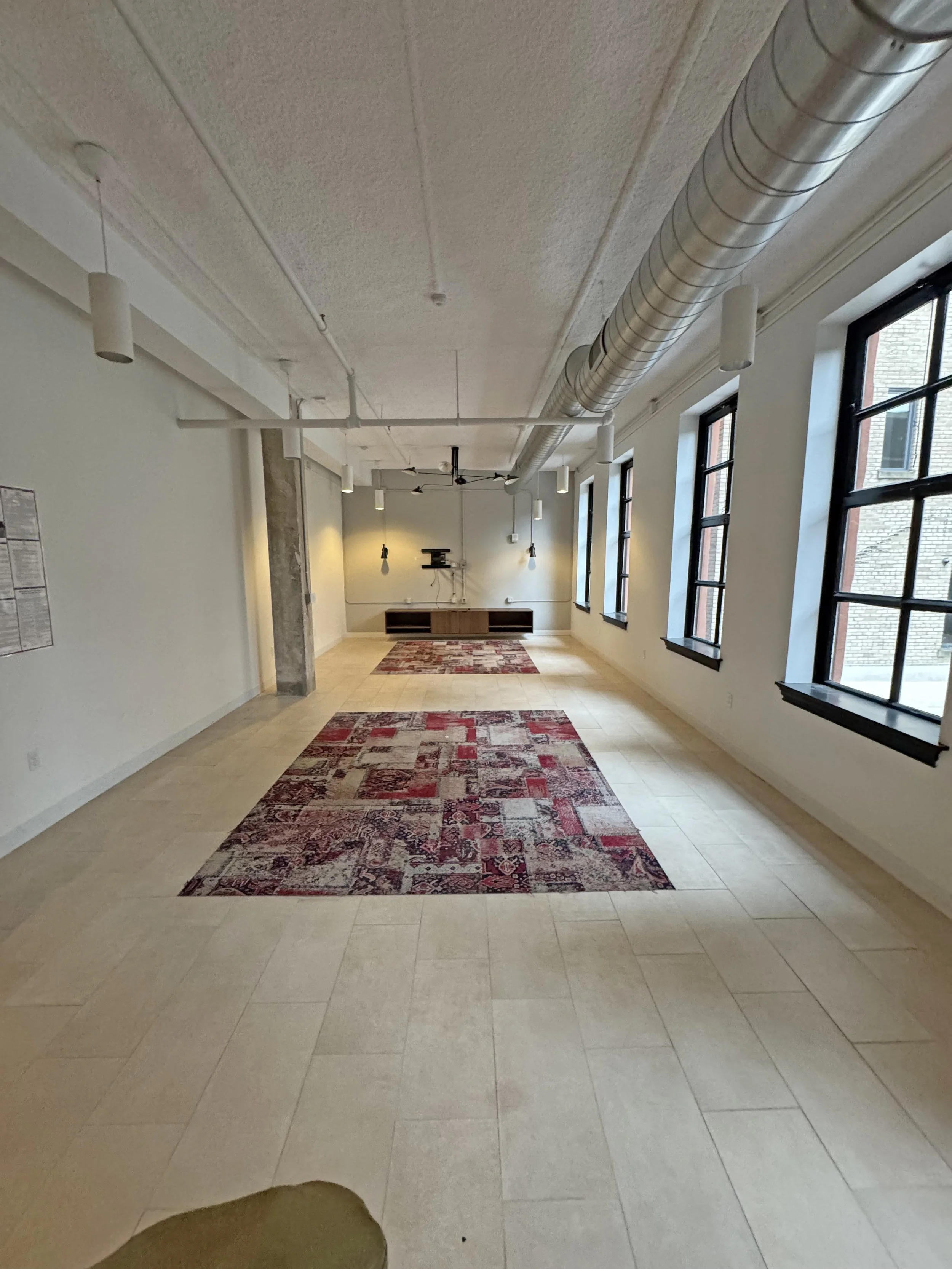 Empty room with large windows, light-colored tiled floor, patterned red and beige rugs, a low wooden cabinet on the far wall, and exposed ceiling pipes.