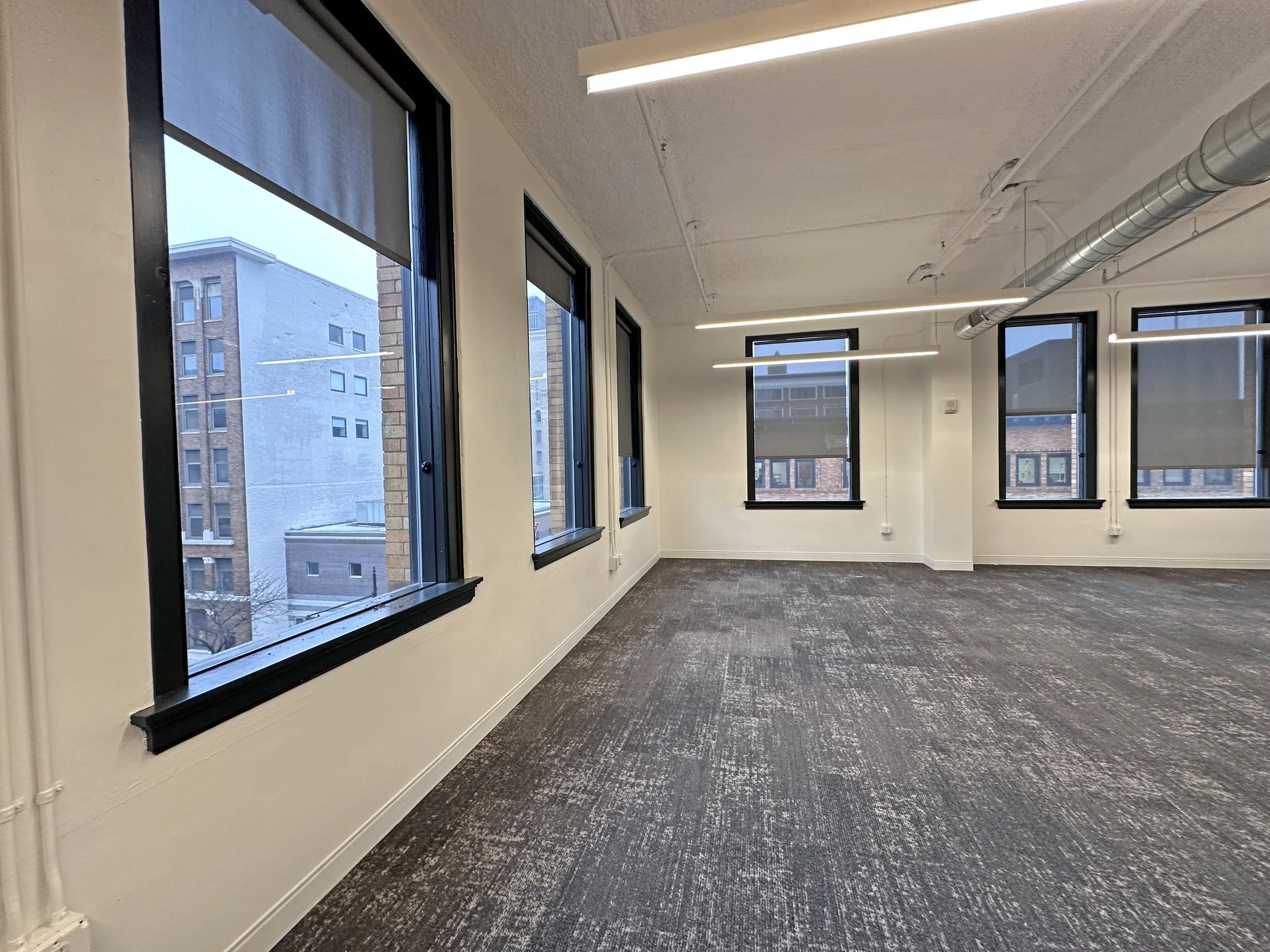 Empty office with large windows, gray carpet, white walls, and exposed ventilation ductwork.
