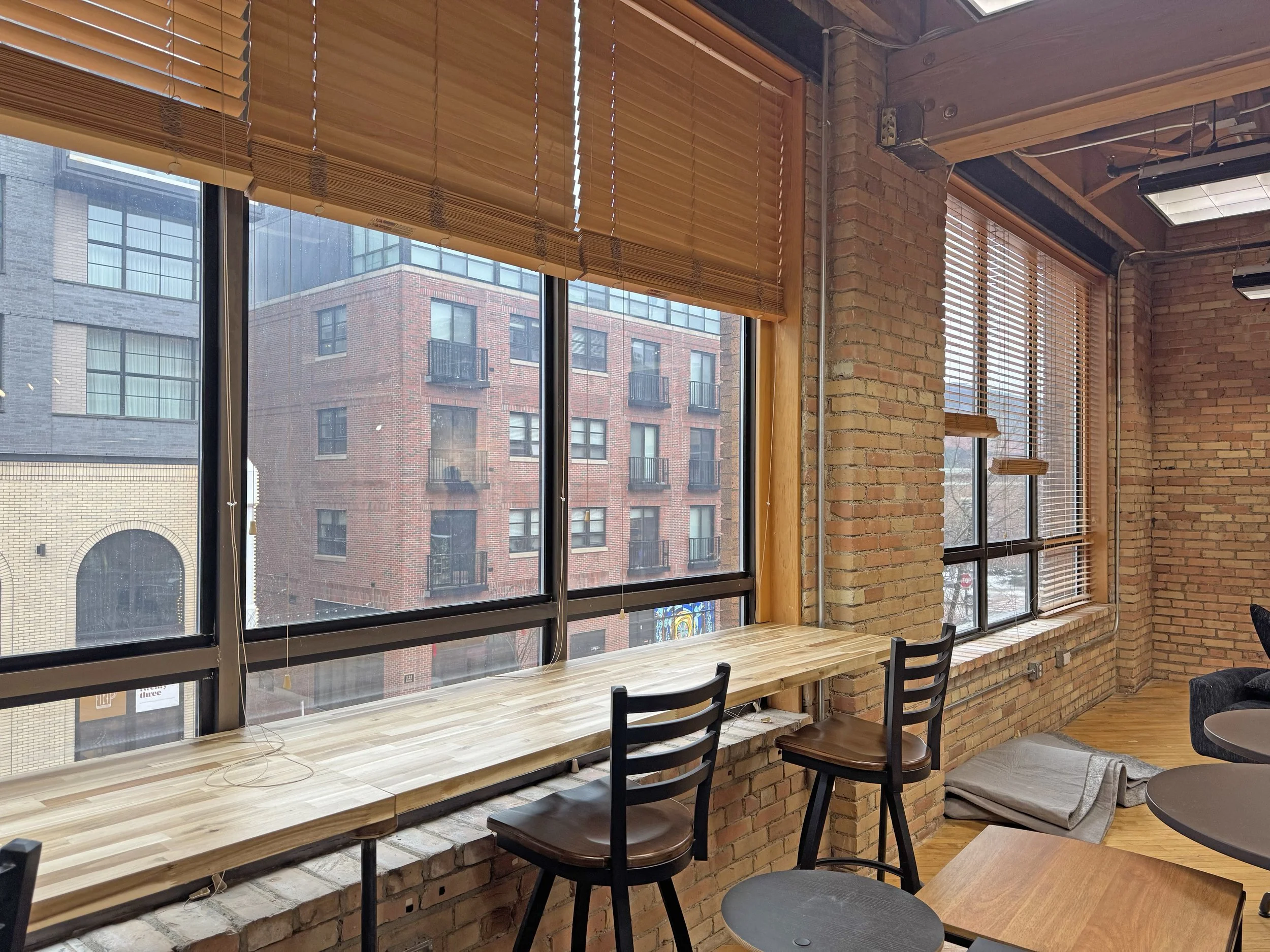 Interior of a modern cafe with large windows, brick walls, wooden blinds, a wooden bar-height table, and black chairs.