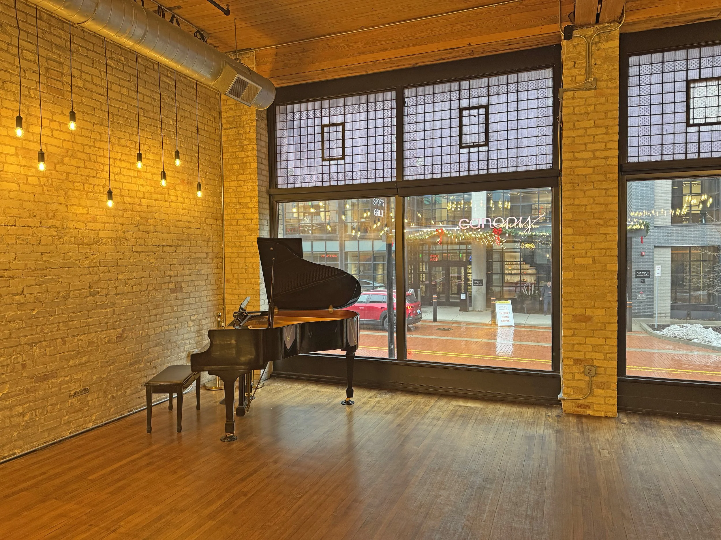 Empty room with a black grand piano and a bench, brick walls, hanging light bulbs, large windows, city street view outside, signs for 'Canopy' and 'Sports Bar' visible in the background.