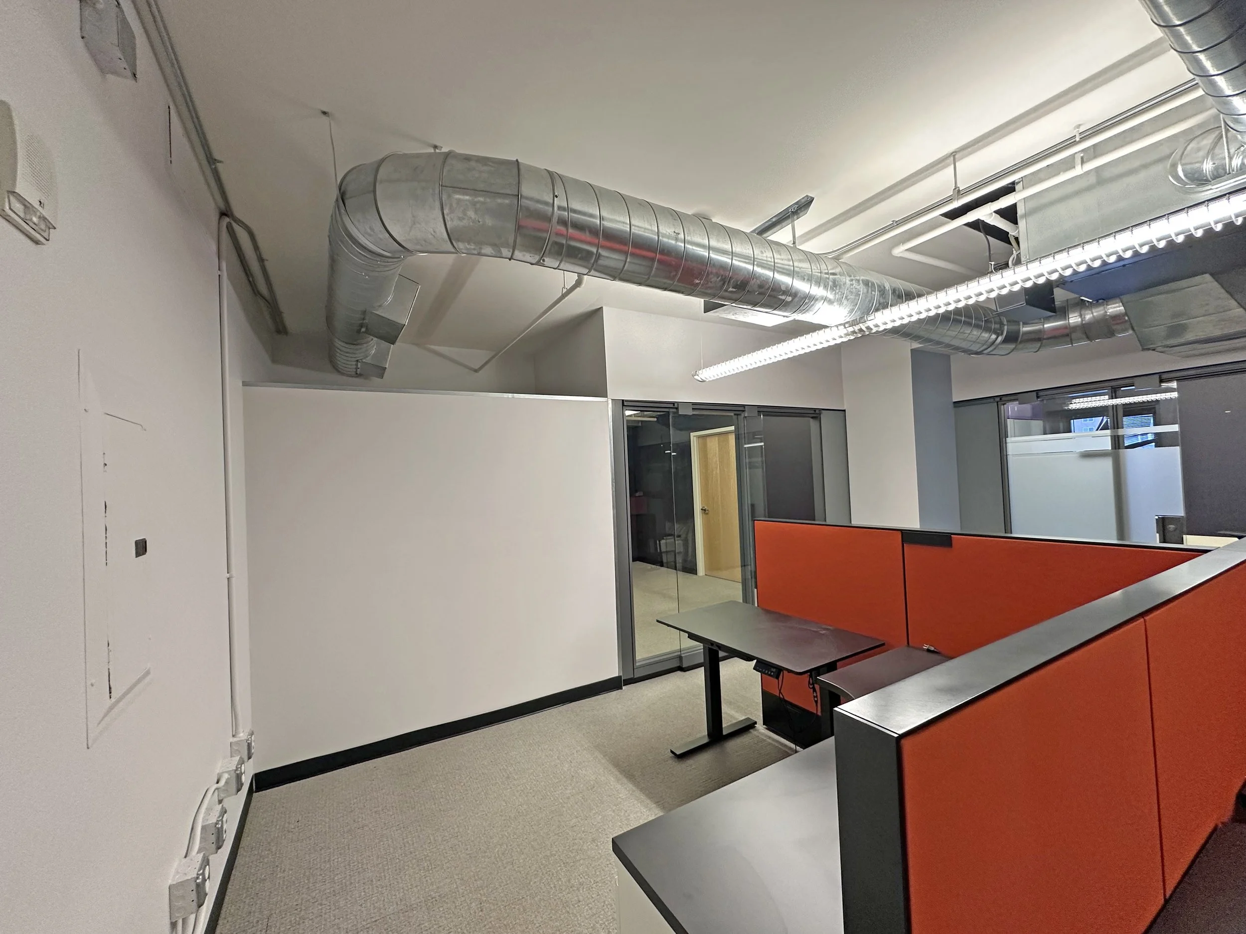 Office space with orange cubicles, a small desk, and visible metallic ductwork on the ceiling.
