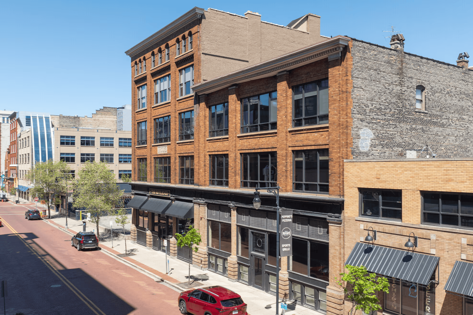 Street scene in a downtown area with multi-story brick buildings, shops, trees, parked cars, and clear blue sky.