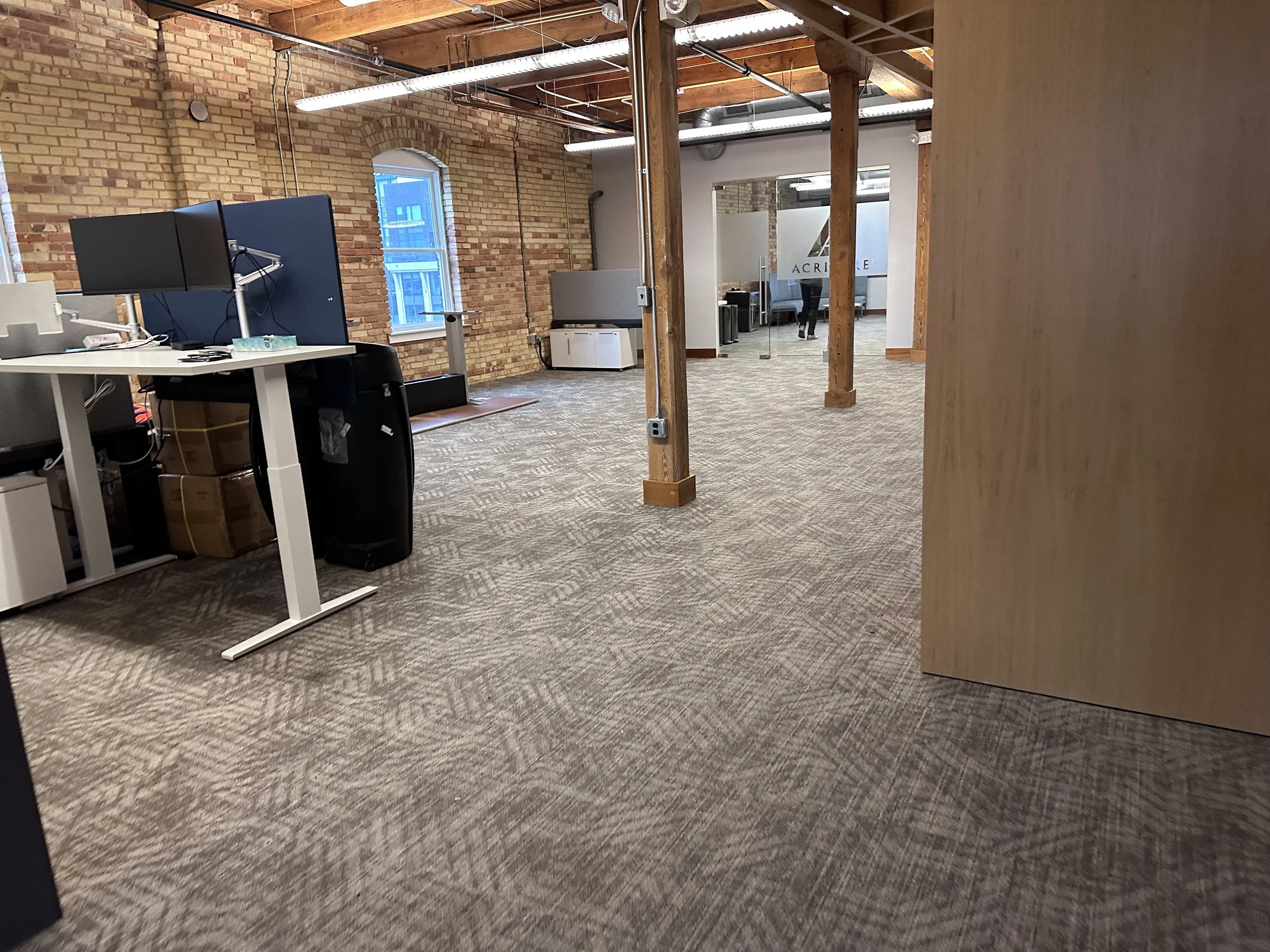 An open office with exposed brick walls, large windows, wooden support beams, patterned carpet, and modern furniture including desks and monitors. In the background, there are glass partitions and a person walking.