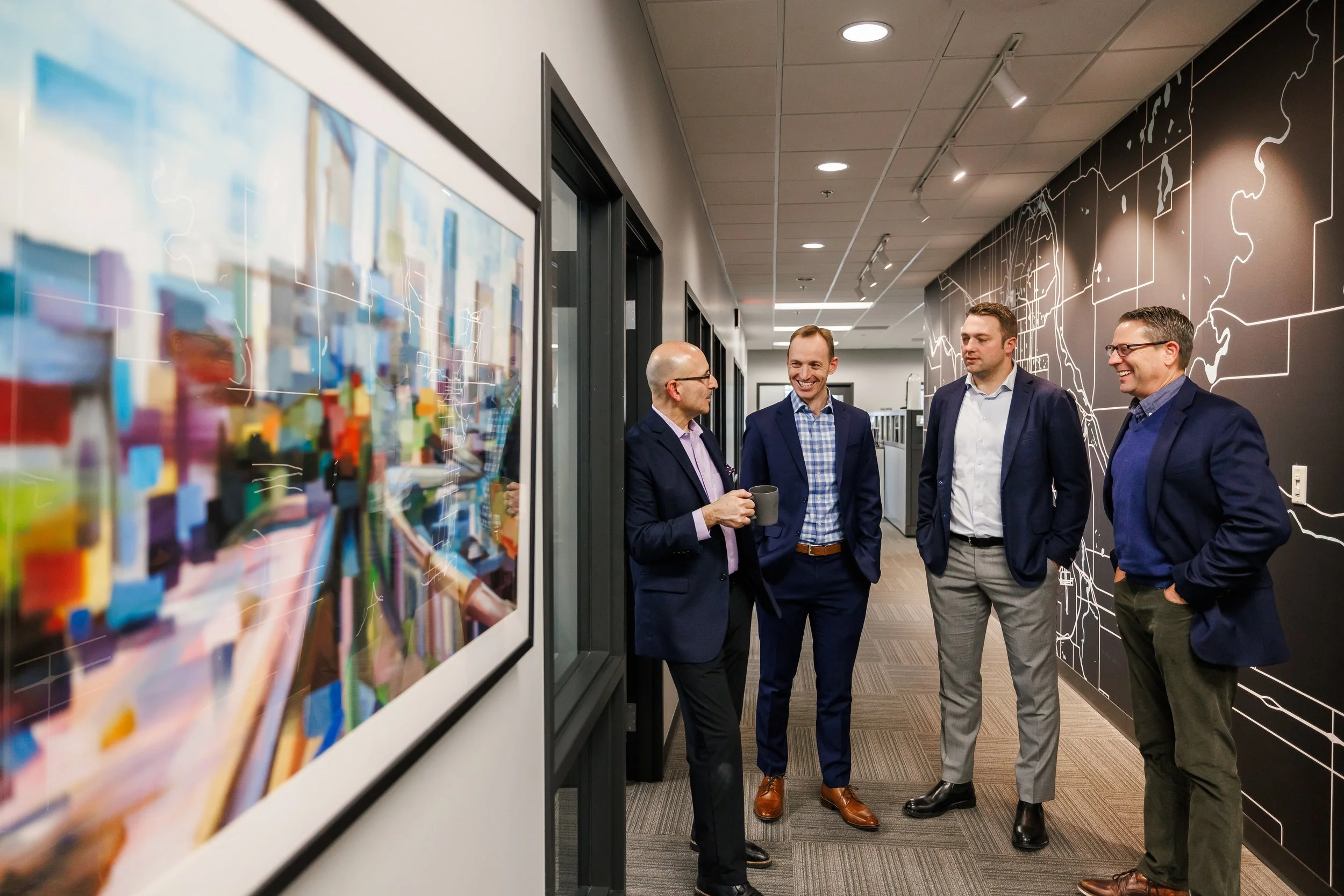 Four Advantage Commercial Real Estate brokers in business attire having a conversation in an office hallway, with one holding a coffee mug, near framed artwork and a large wall map.