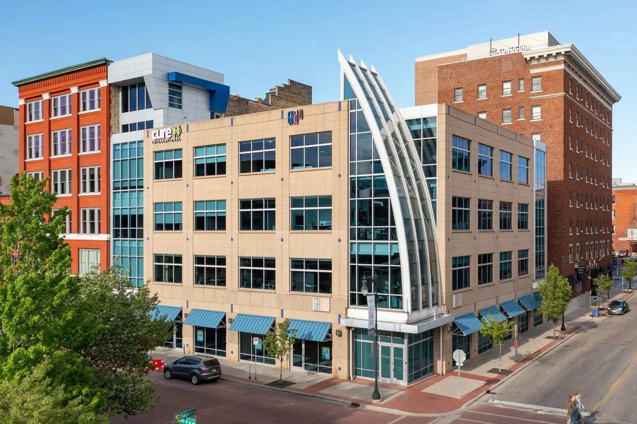 City street view featuring a modern multi-story building with a curved glass entrance, adjacent traditional brick buildings, parked cars, and pedestrians.