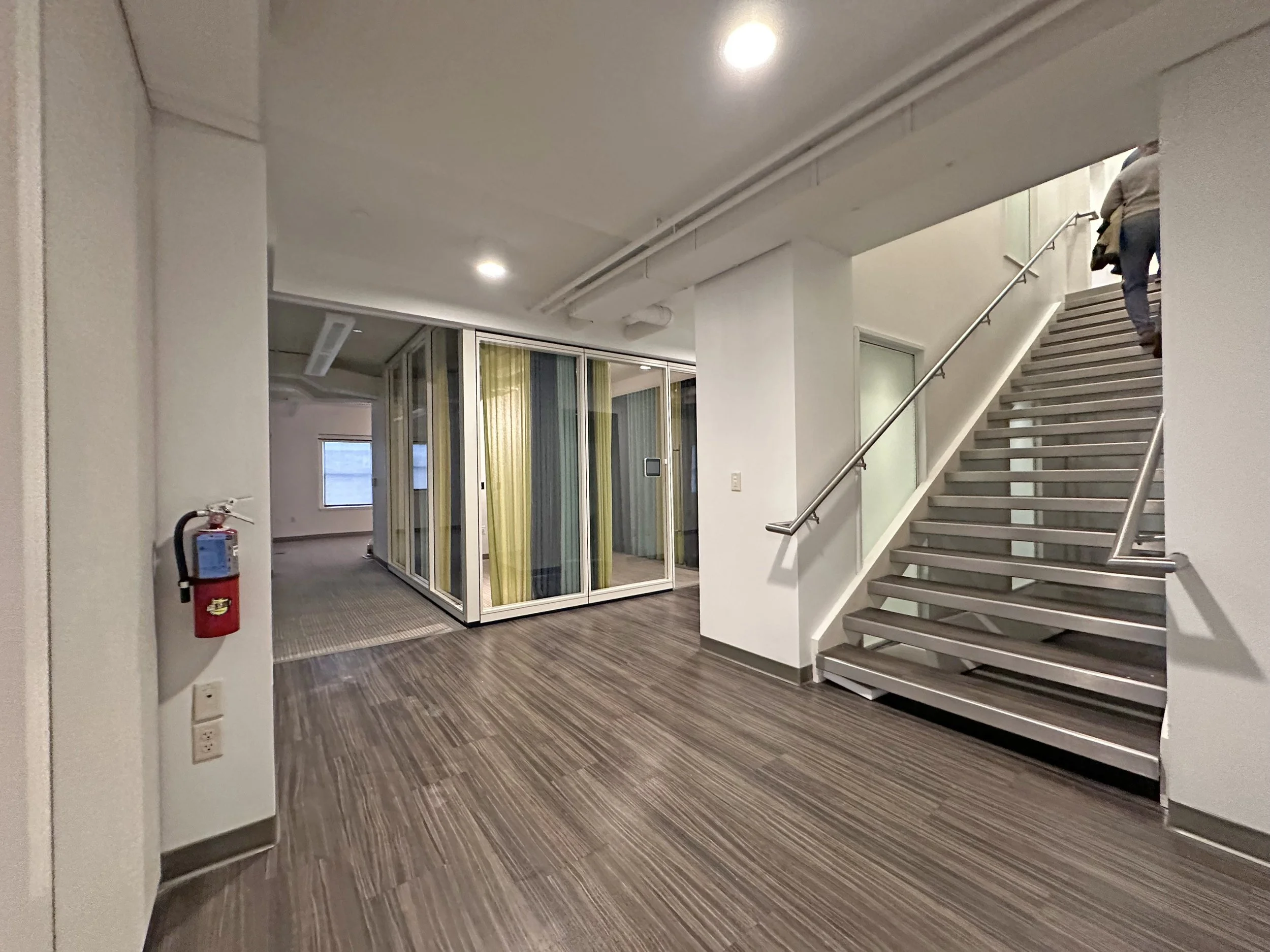 Interior view of a modern building staircase with metal stairs and glass panels, a fire extinguisher on the wall, and an enclosed office space with colorful curtains in the background.