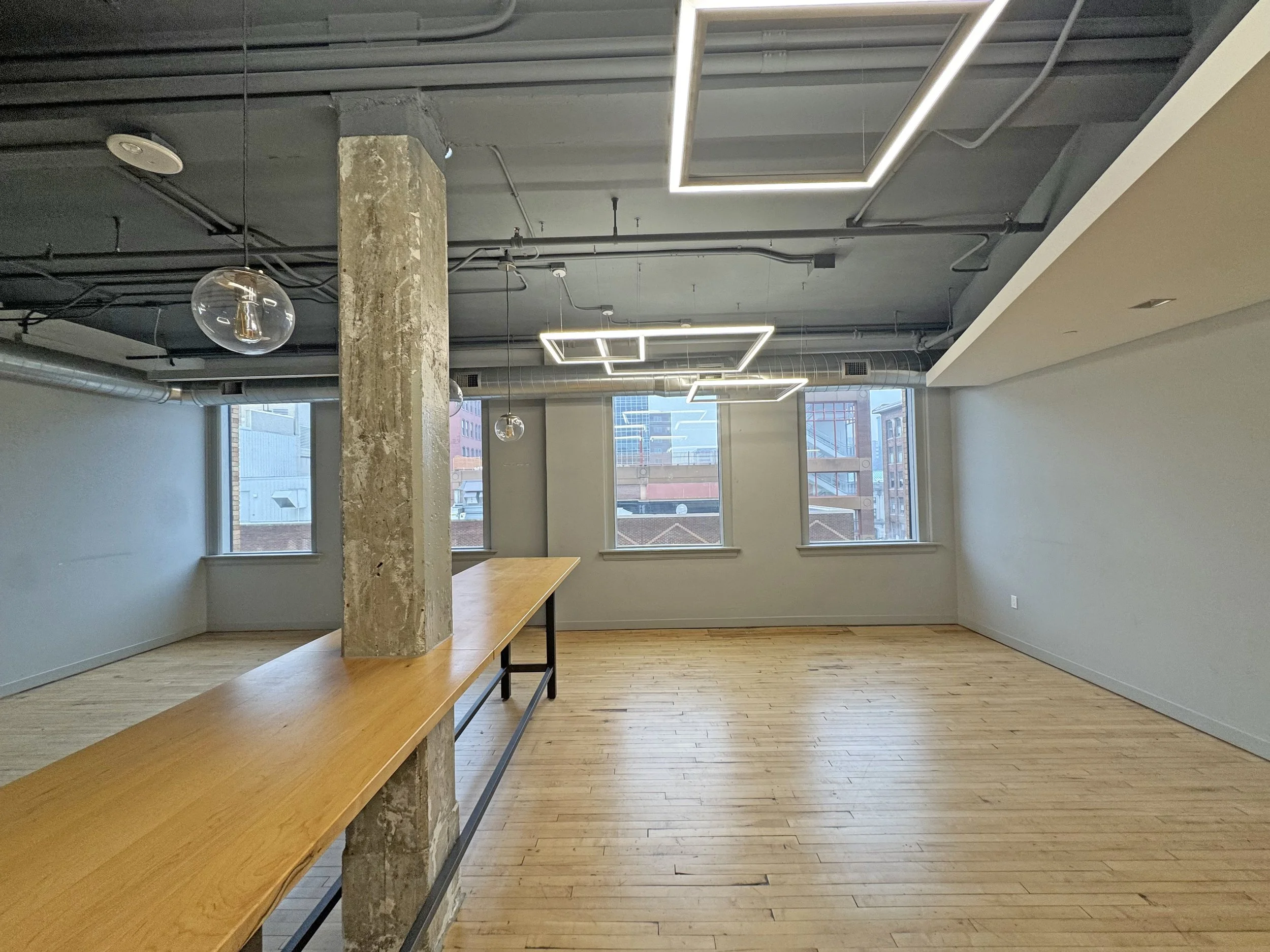 Empty modern office space with wooden floors, large windows, exposed ceiling with lighting fixtures, and a long wooden table supported by a concrete pillar.