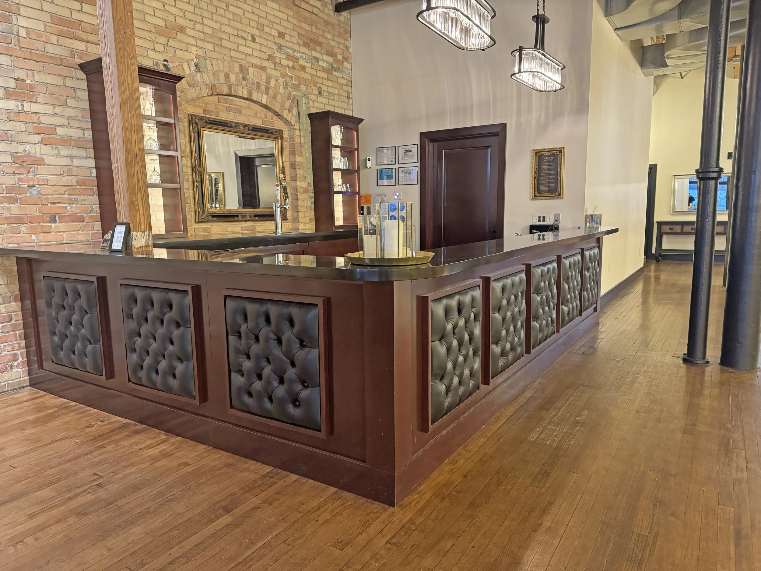 A hotel or restaurant reception desk with dark wood and tufted leather panels, a brick wall, a mirror, decorative glass light fixtures, and hardwood flooring.