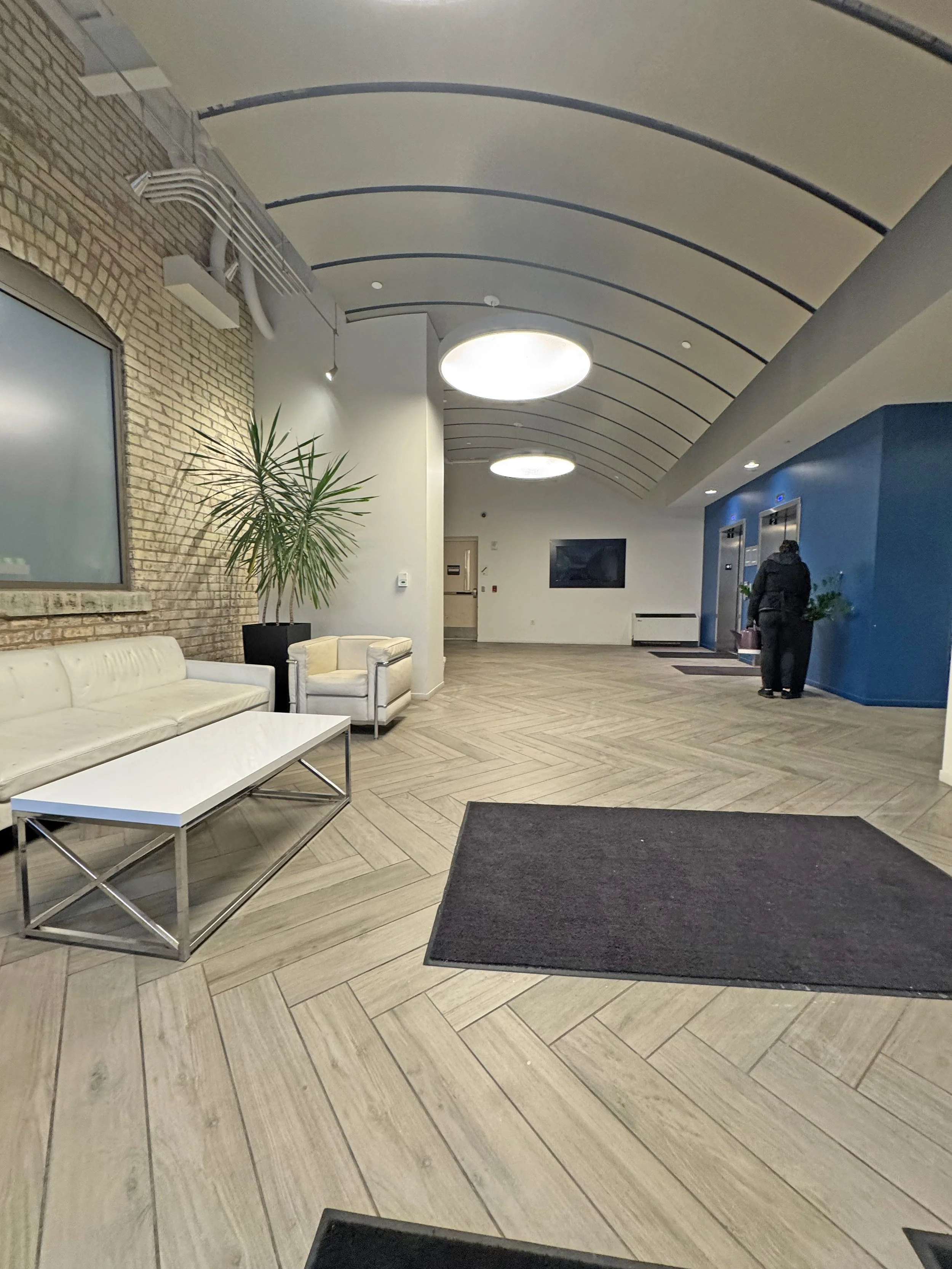 Modern hotel lobby with two white sofas, an indoor plant, a large flat-screen TV on the wall, and an elevator with two people standing nearby. The ceiling has curved lines with recessed lighting, and the floor is covered with light wood-patterned tiles.