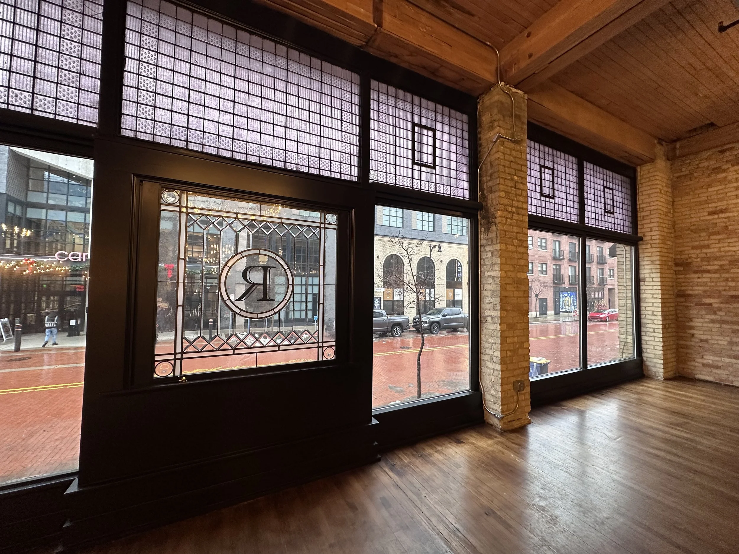 Empty interior space with large front windows showing an urban street scene outside, brick and wood accents, and a logo with the letter 'R' visible on one window.