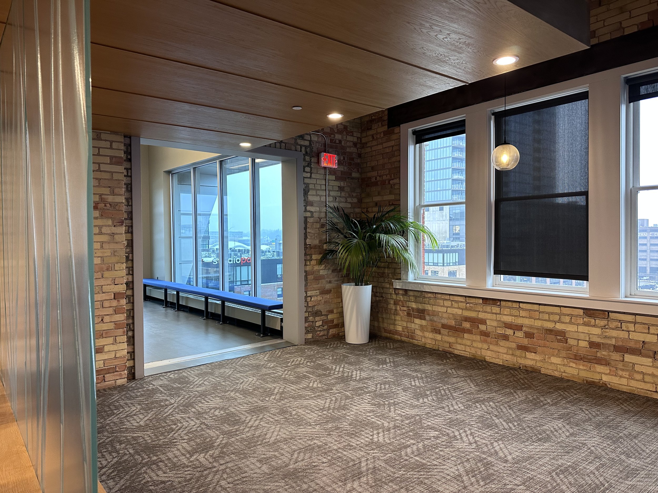 Interior of an office space with brick walls, large windows with black shades, a potted plant, and a view of city buildings outside.