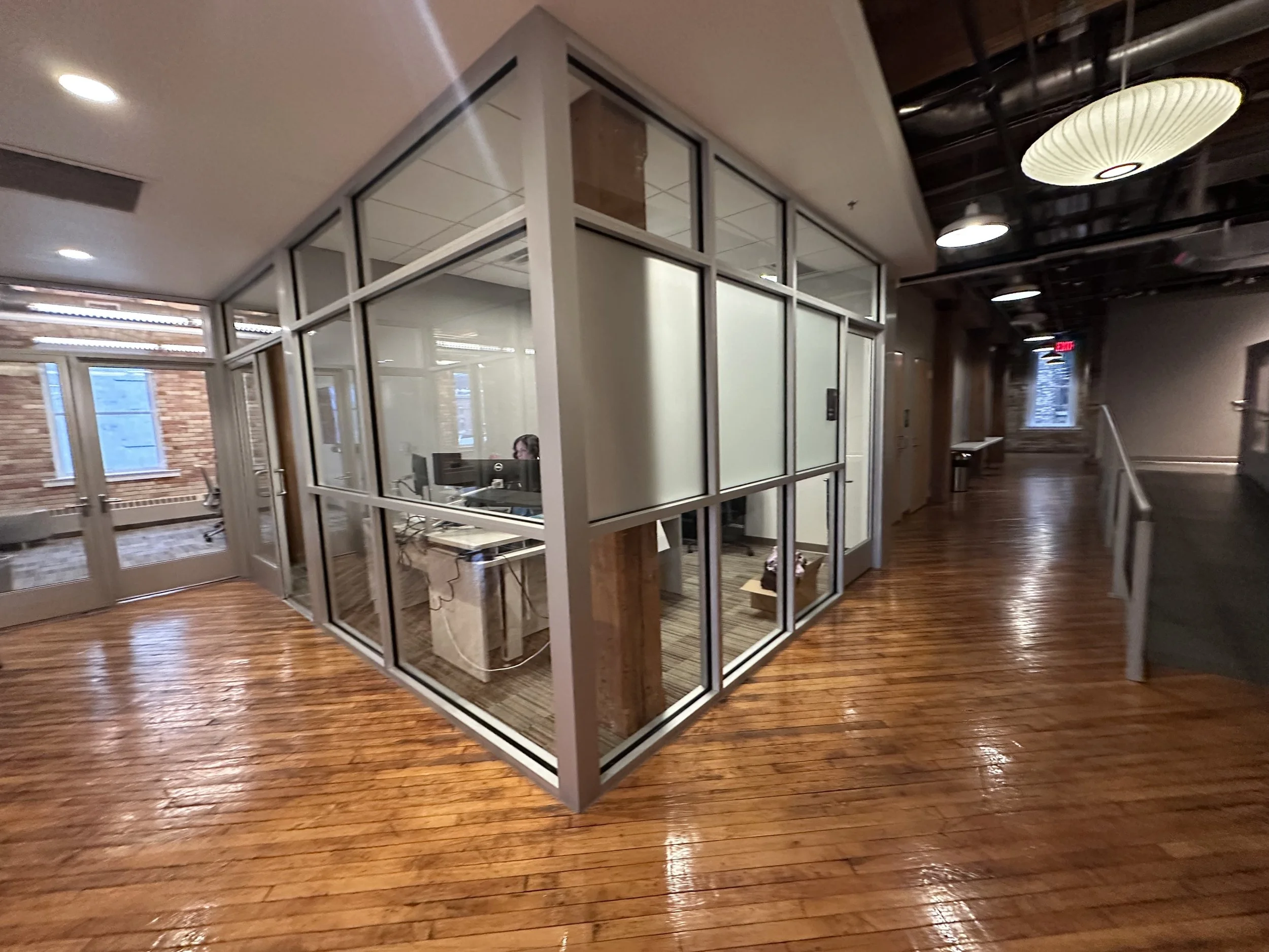 Interior view of an office space with a glass-paneled room, wooden floors, and a hallway with lighting fixtures.