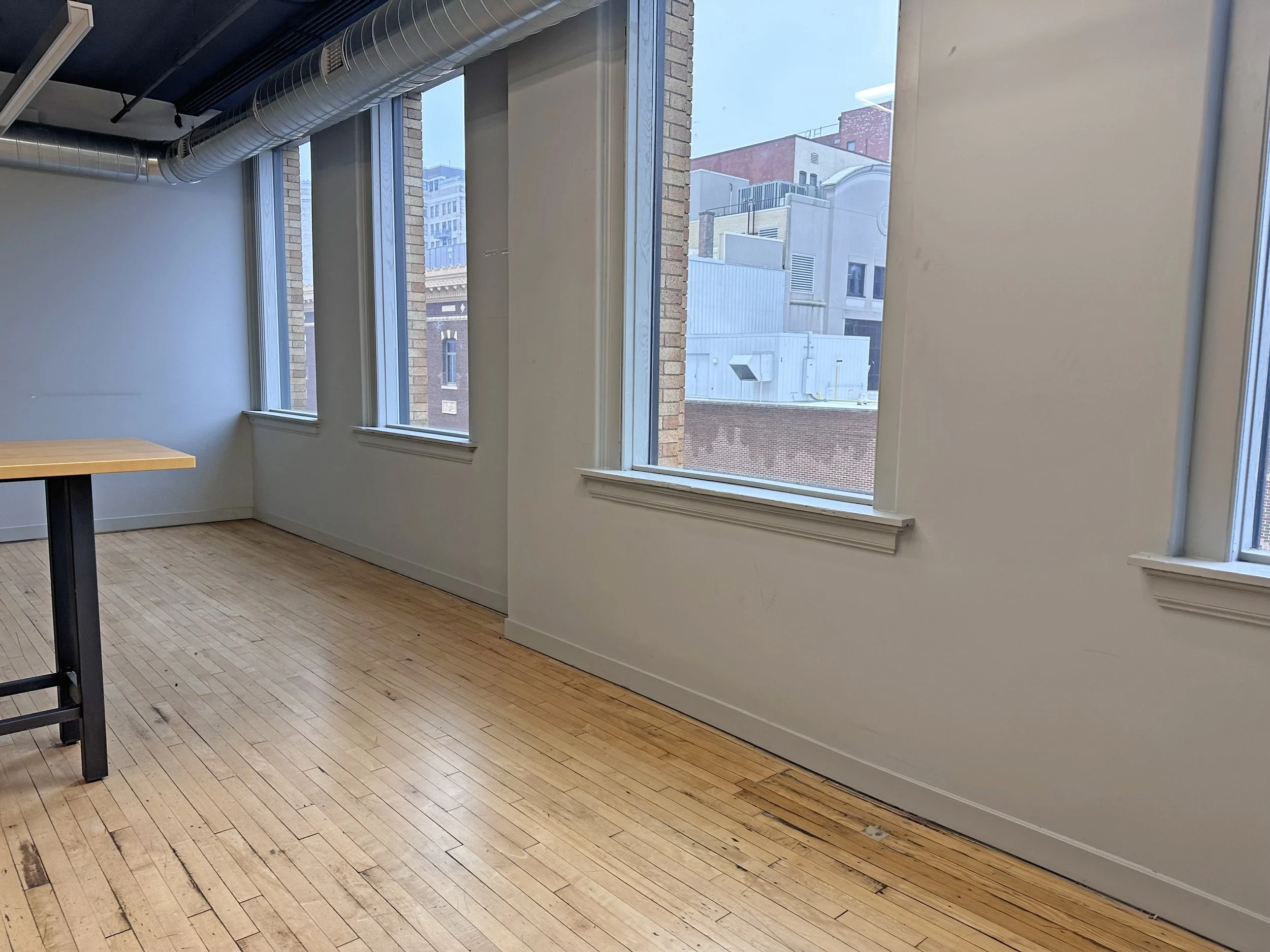 An empty room with three large windows, white walls, wooden flooring, and a black table leg in the corner.