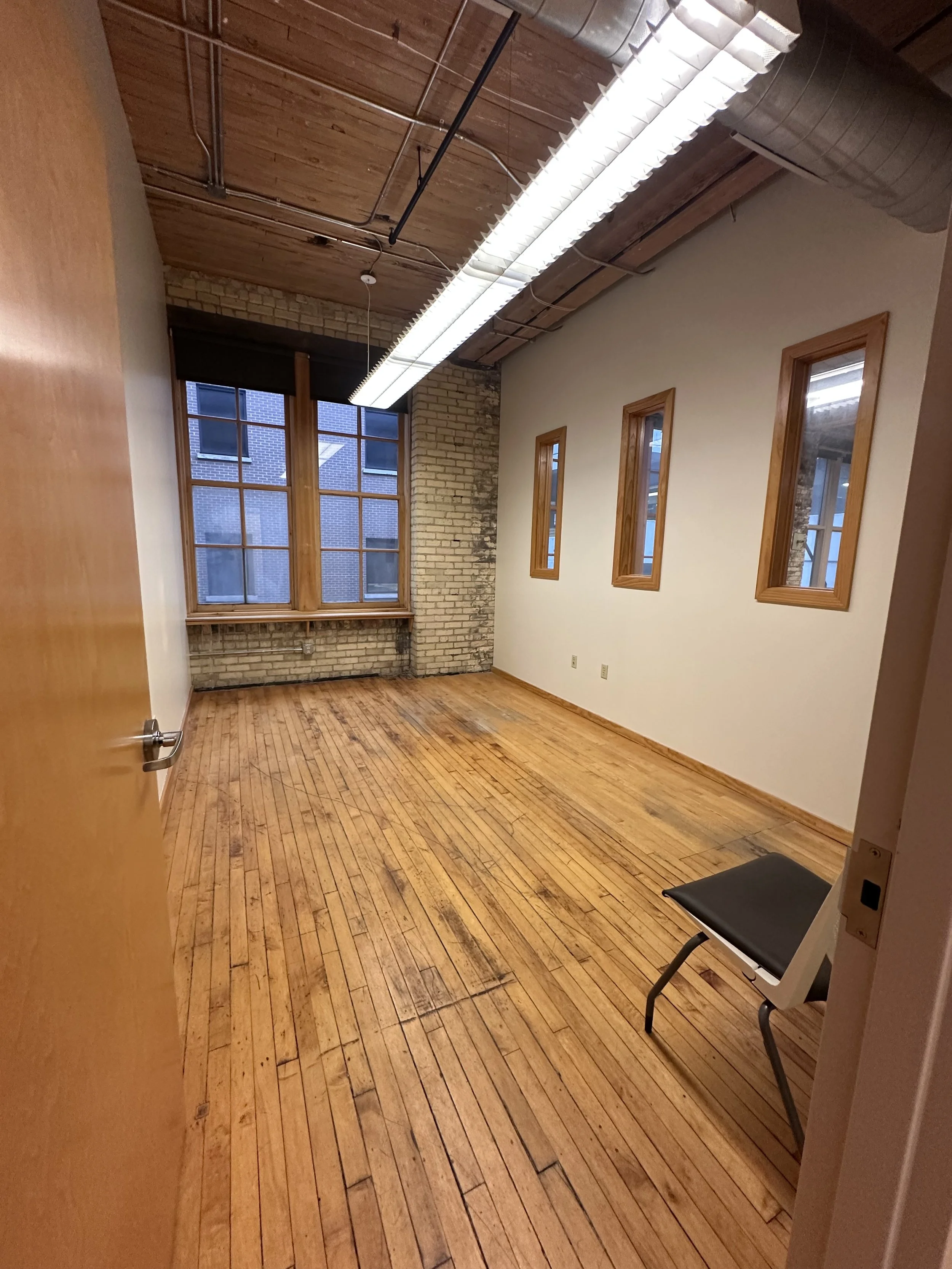 Empty office room with wooden floors, large windows, brick wall, wooden framed windows, and a black chair with gray frame.