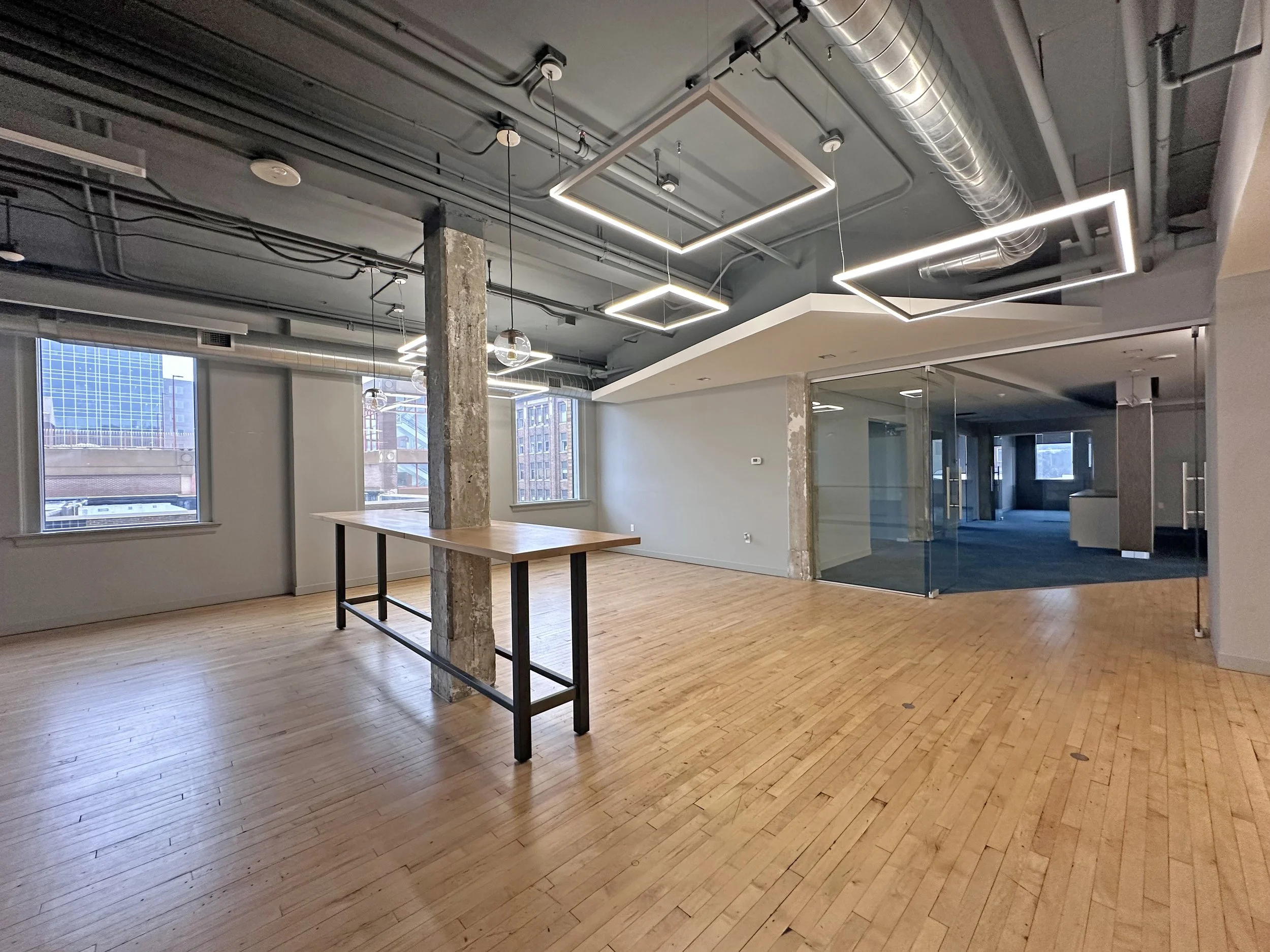 Empty modern office space with wooden floors, large windows, exposed ceiling pipes, and geometric light fixtures. There is a wooden table positioned near a concrete pillar and glass-walled rooms in the background.