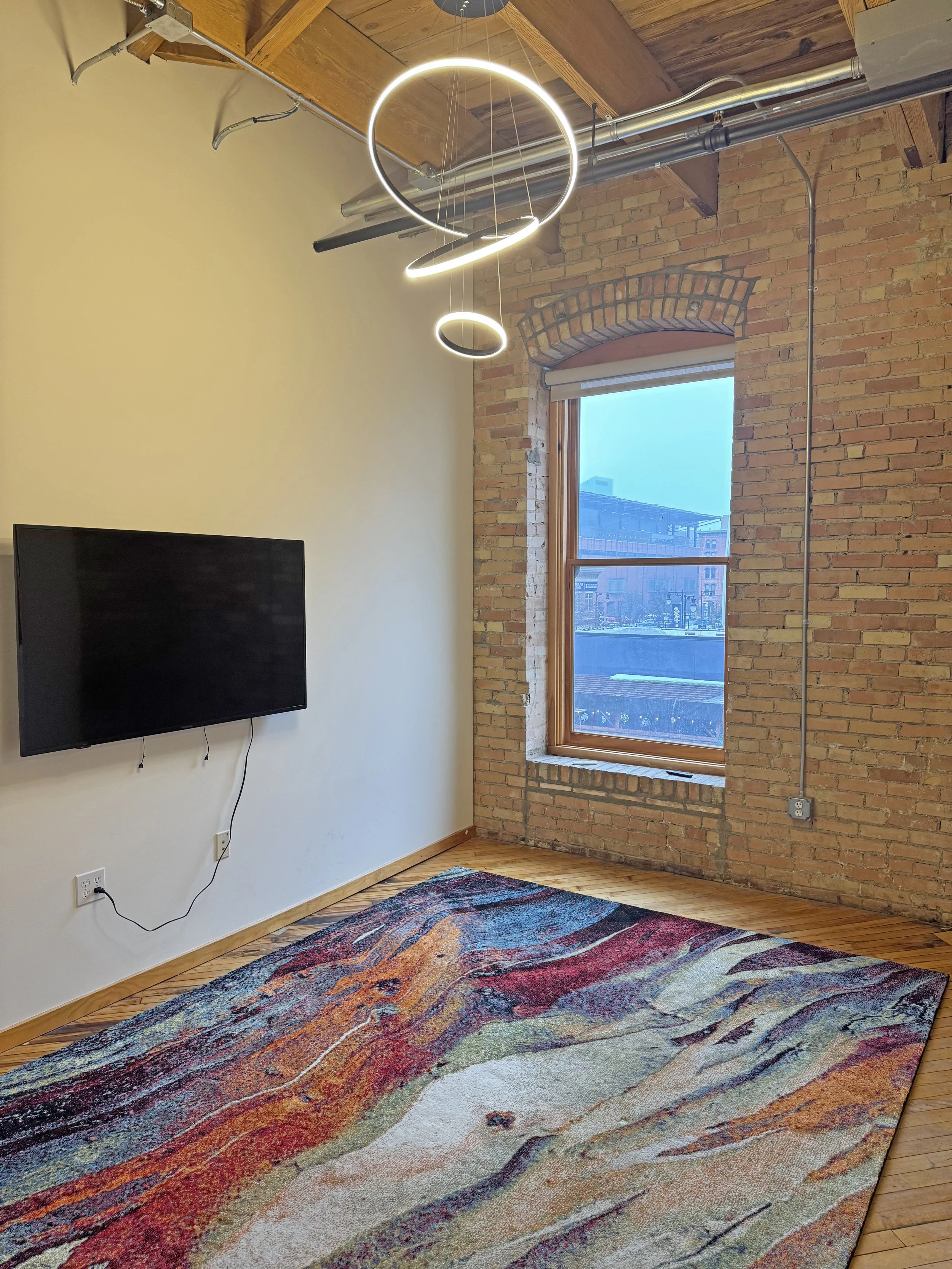 Modern interior room with an exposed brick wall and wooden ceiling, featuring a circular ceiling light and a window, with a colorful abstract area rug and a mounted flat-screen TV.