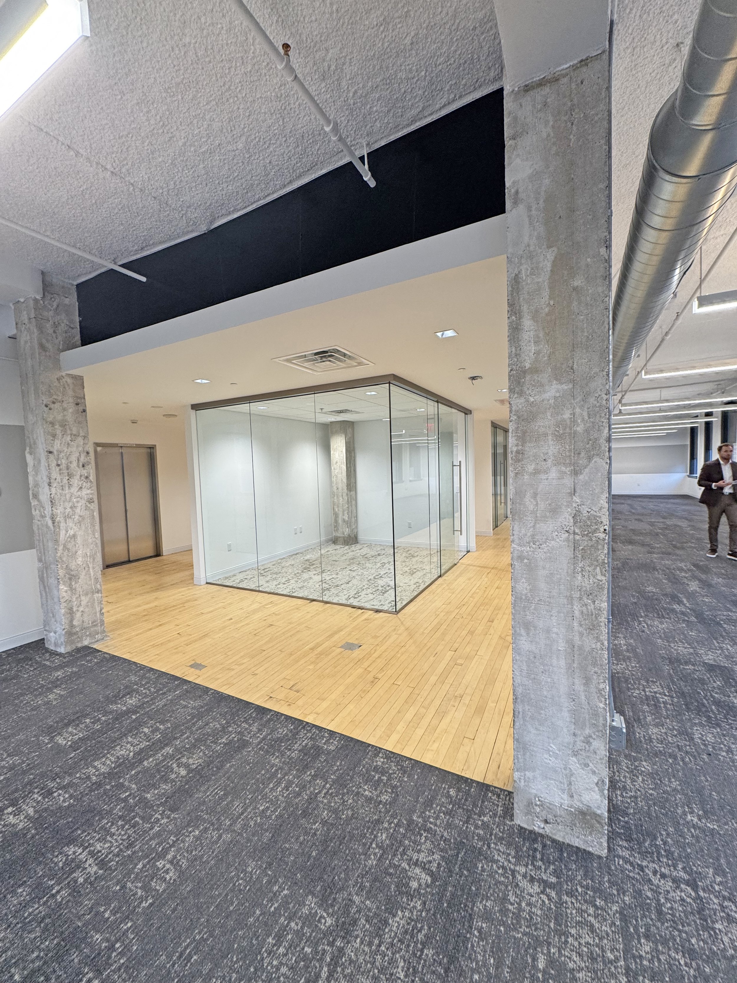 An empty modern office space with a glass-walled conference room, wood flooring, and an industrial-style ceiling with exposed ductwork.