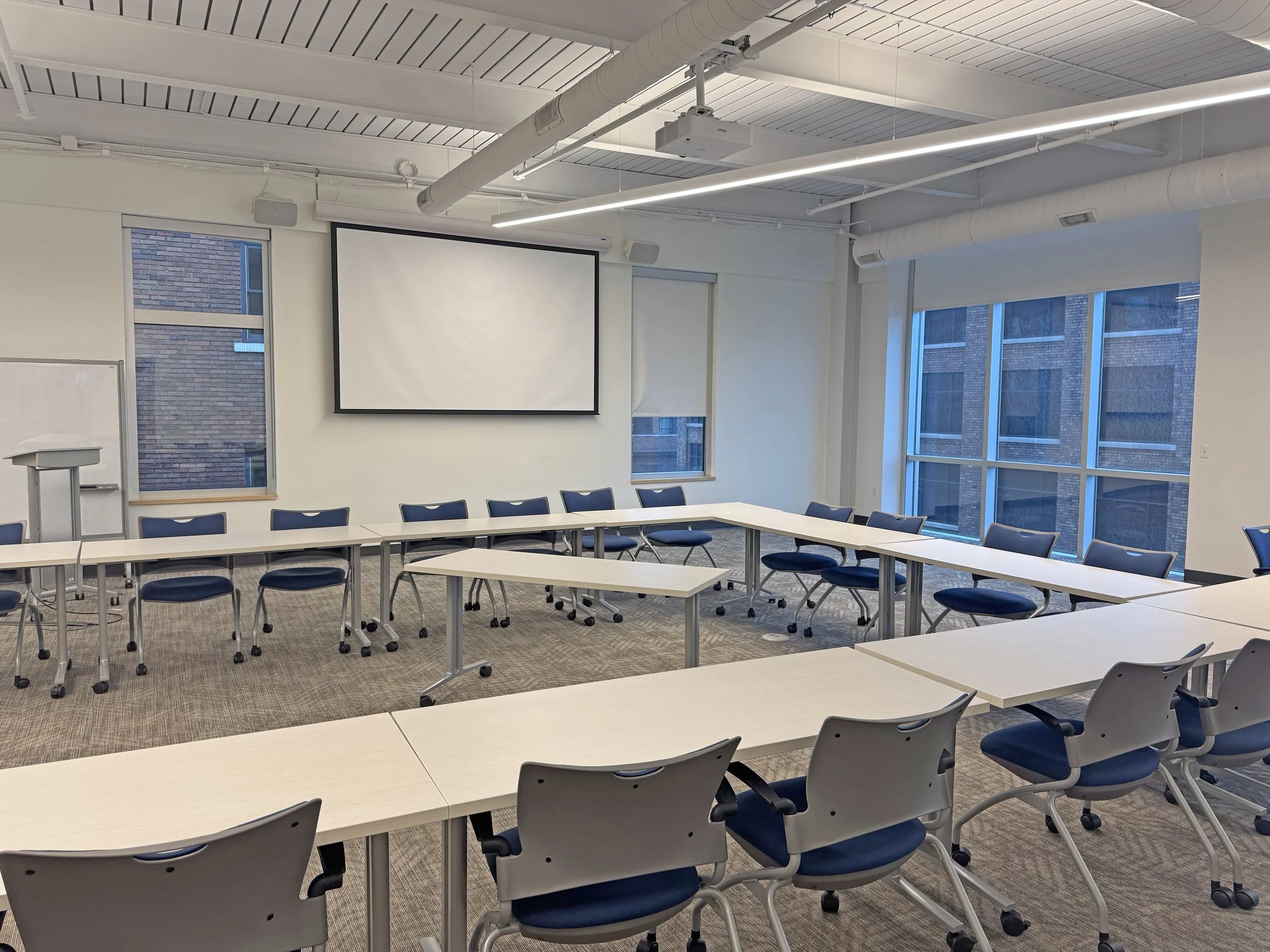 Empty conference room with U-shaped arrangement of white tables and dark blue chairs, a large white projection screen on the wall, windows with brick buildings outside, and a whiteboard to the side.