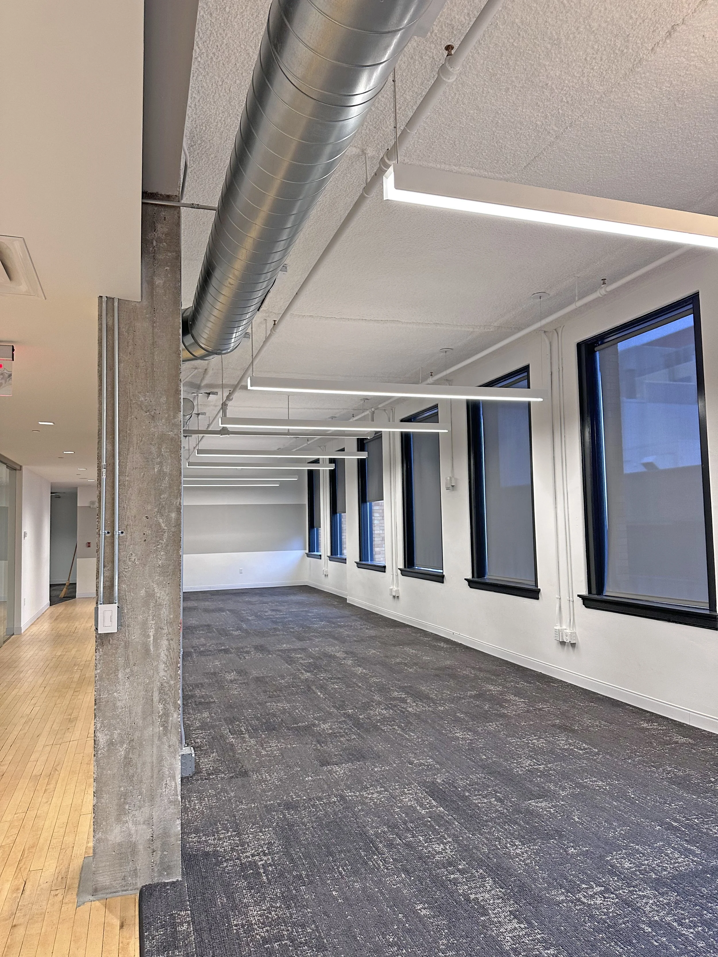 A modern office space with multiple large windows, ceiling-mounted linear LED lights, and an exposed ductwork on the ceiling. The room has a combination of carpeted and wooden flooring.
