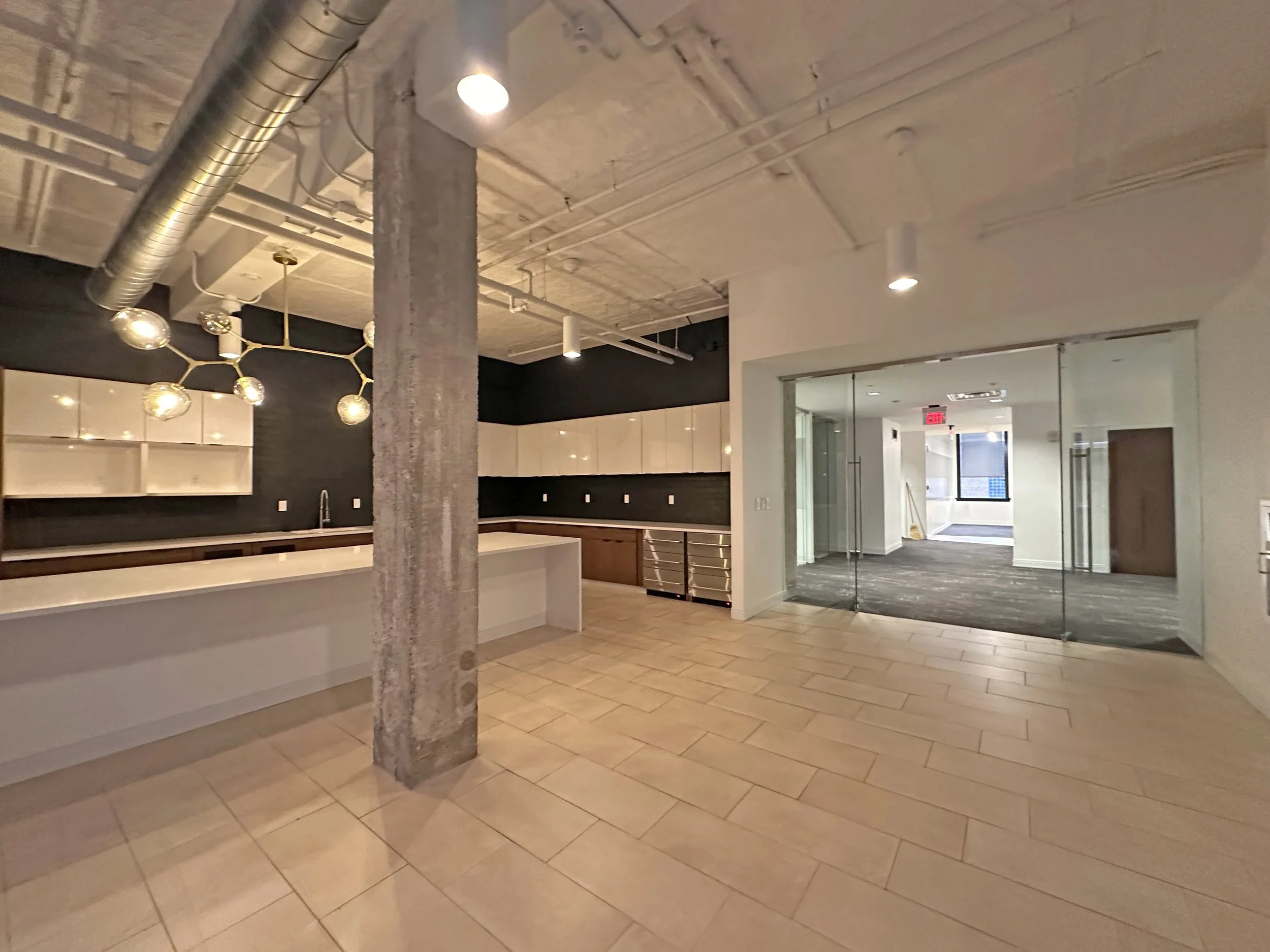 Empty modern commercial kitchen with beige tile flooring, black and white cabinetry, a large kitchen island, exposed concrete column, industrial ductwork, and a glass door leading to a carpeted room with windows.