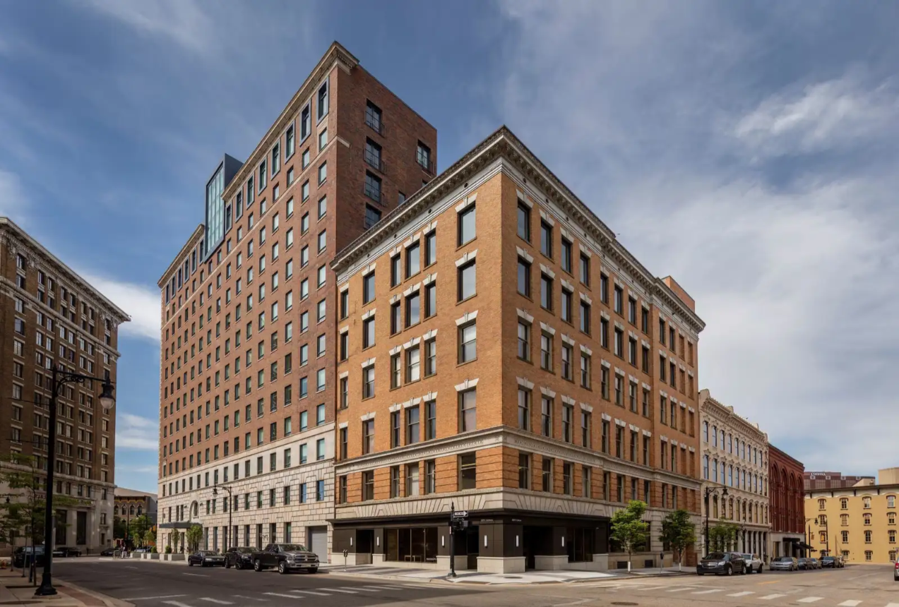Tall multi-story brick buildings on a city street with parked cars and trees.