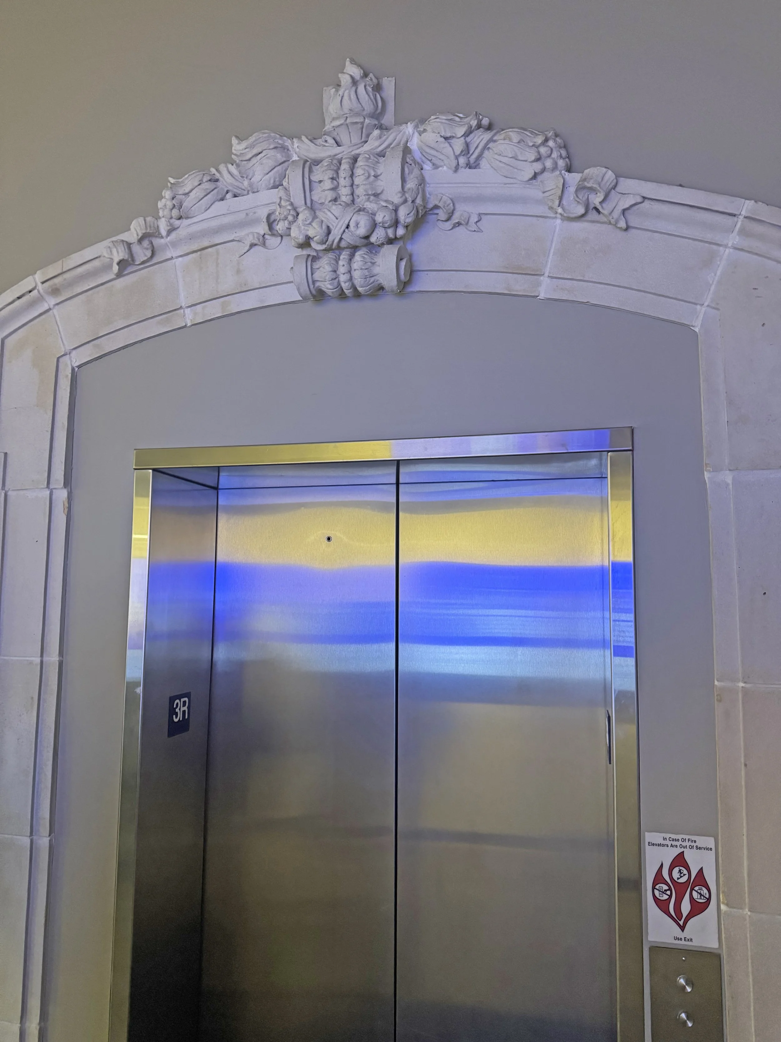 Decorative architectural detail above elevator door with ornate stone carvings, and a stainless steel elevator door with a small black sign reading '3R' and a fire safety sticker.