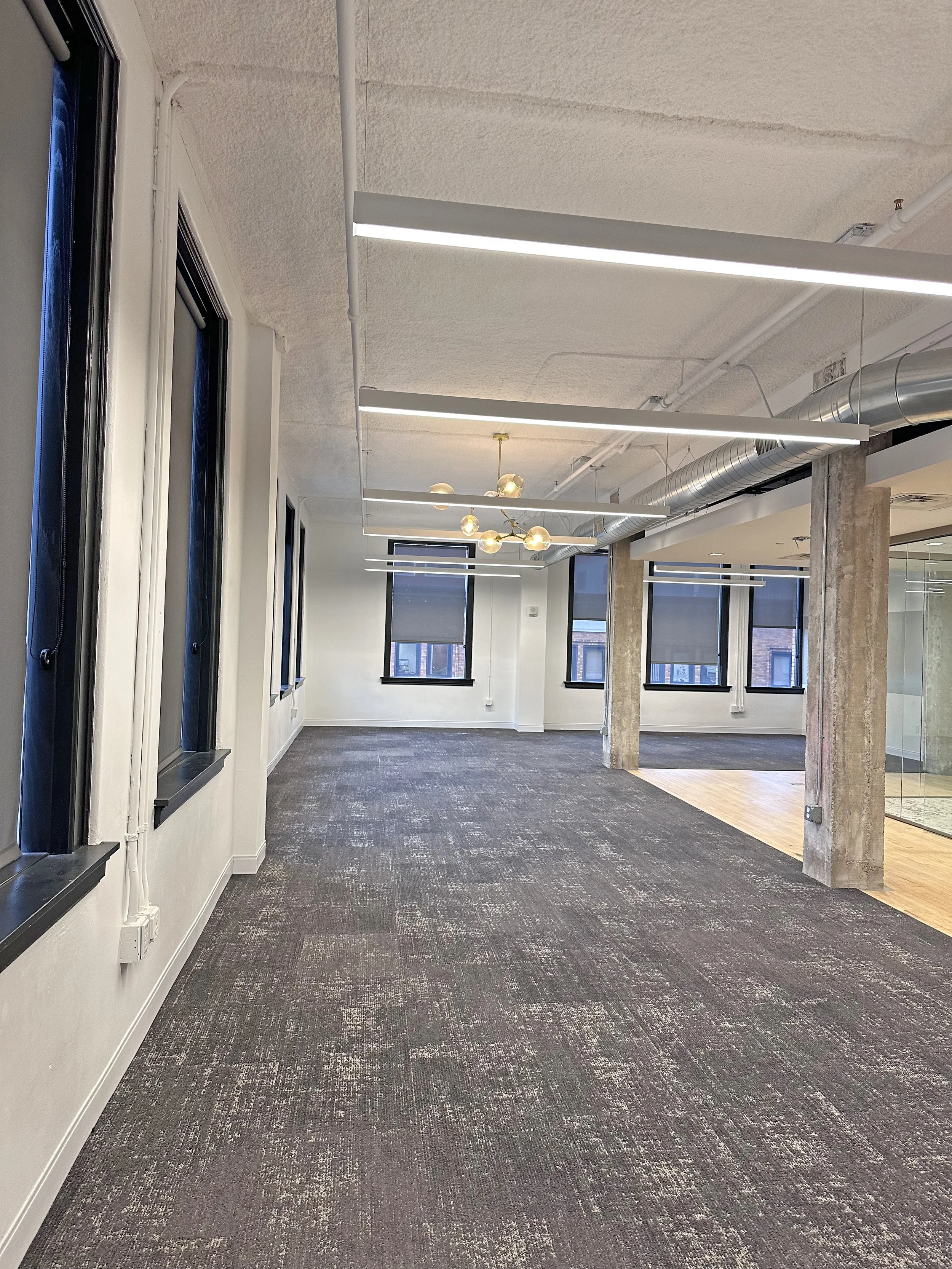 Empty modern office space with large windows, carpeted floors, exposed concrete pillars, and ceiling light fixtures.