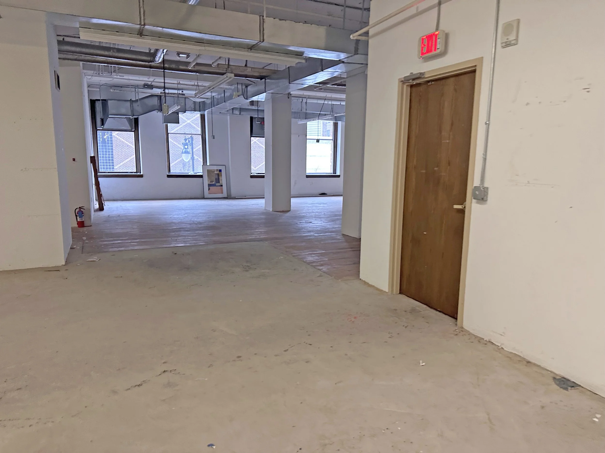 Empty room under construction with exposed ceiling pipes and ducts, wooden floors, and large windows, seen from a foreground with unfinished flooring, and a red exit sign above a wooden door.