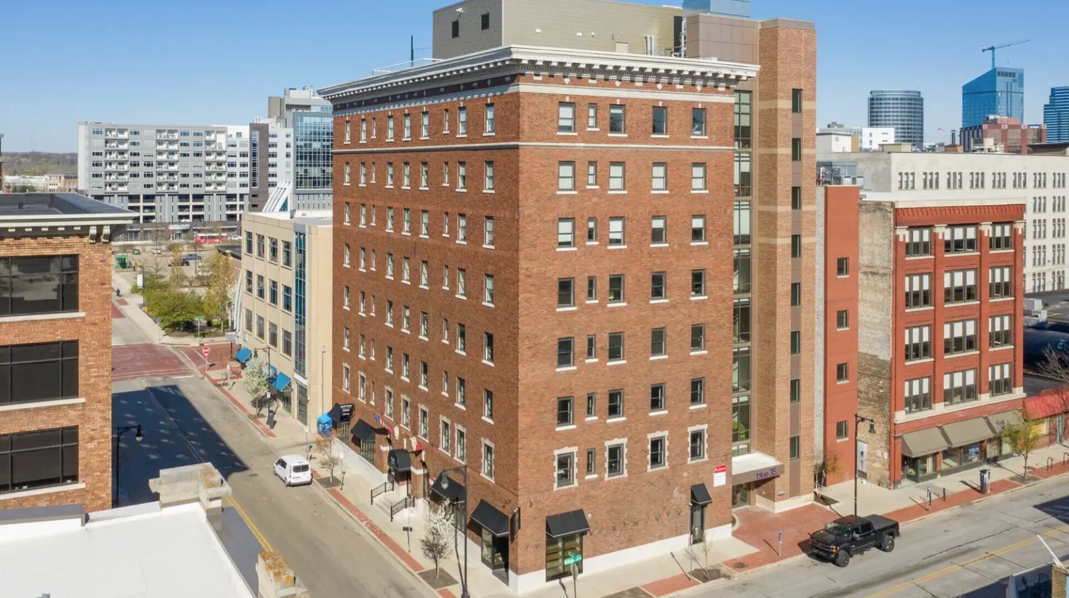 View of a city block with a tall brick building at the corner, surrounded by other mid-rise buildings, with a street and sidewalk below. The scene is under clear blue skies.