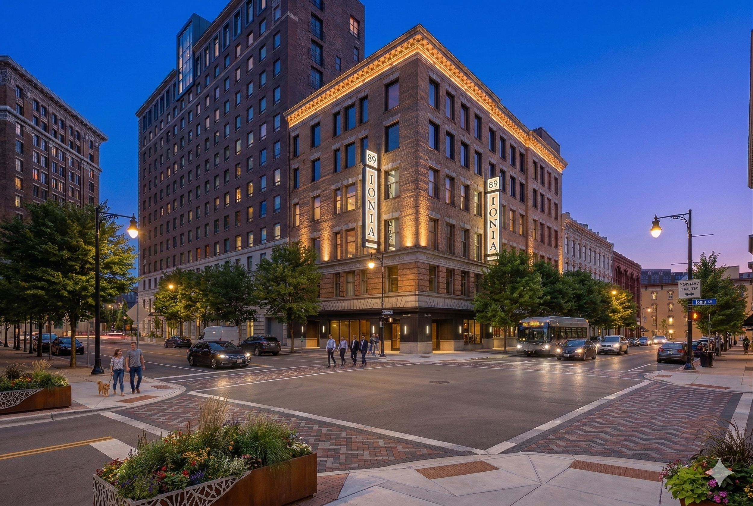 Grand Rapids street scene at dusk with a large, illuminated brick building named IONIA at the corner, surrounded by other buildings, trees, streetlights, and cars, with pedestrians walking on the crosswalks.