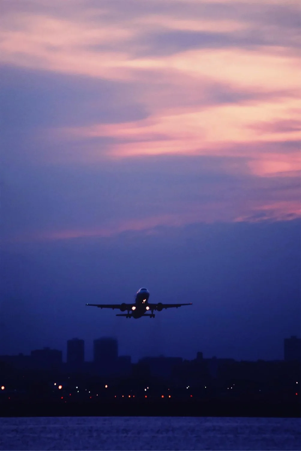 An airplane flying low over a city skyline during sunset with pink, purple, and blue sky clouds.