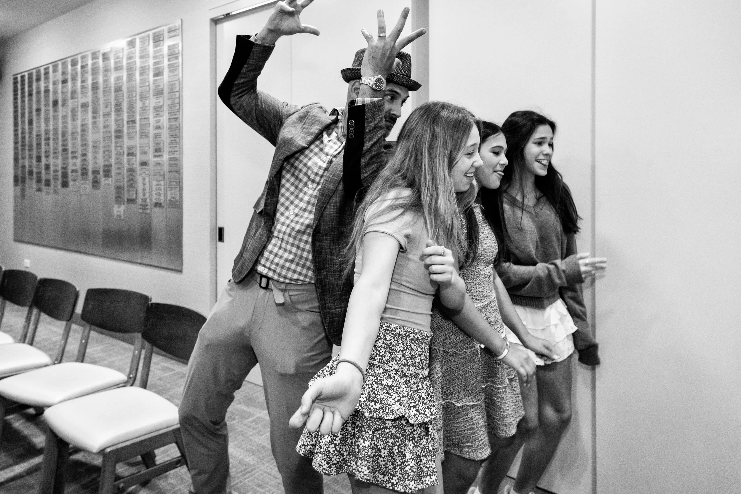 A man and four young women standing in a row, holding hands, with the man raising his other hand in a wave. The scene appears to be in a conference or meeting room, with chairs and a wall with charts or posters in the background.