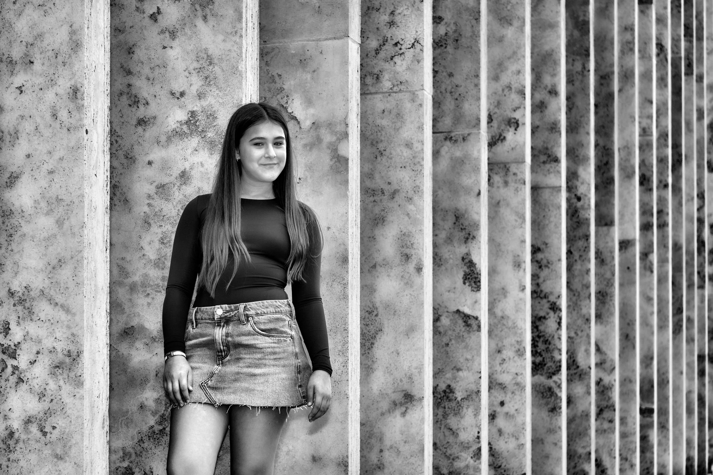 A young woman with long hair wearing a long-sleeved black shirt and denim shorts, standing outdoors against a textured concrete wall.