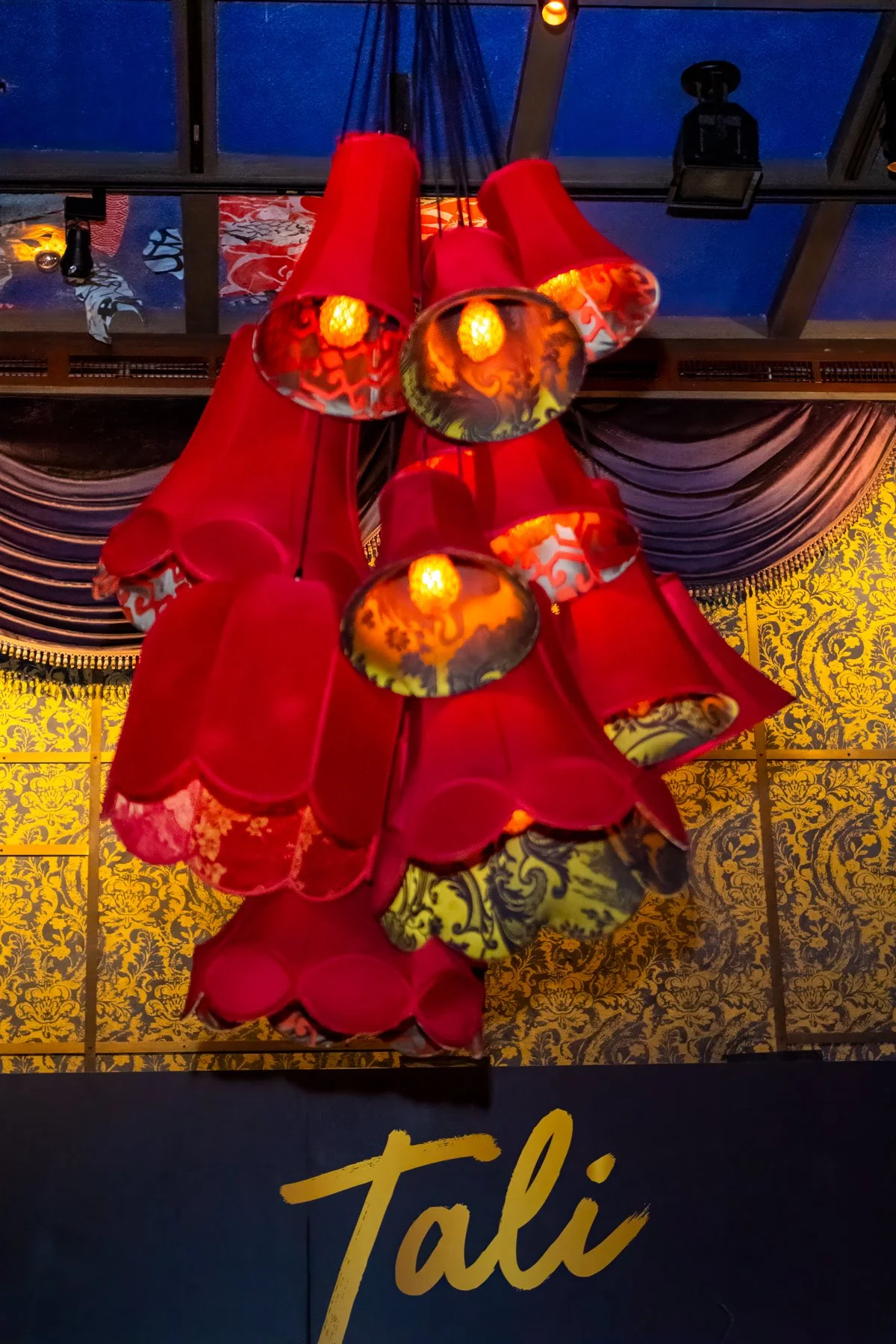 A chandelier with red fabric lampshades and intricate yellow and red patterns reflects on the ceiling. The background features a dark curtain with gold patterns and the word 'Tali' in gold on a dark surface.
