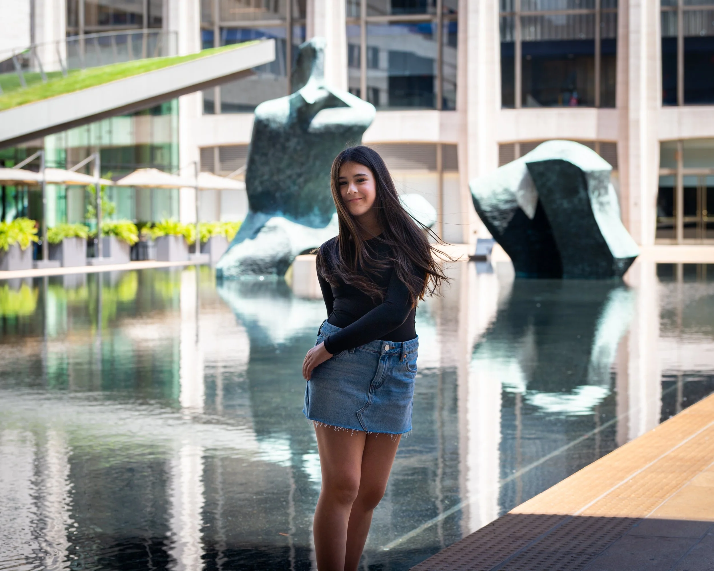 Young woman with long dark hair wearing a black long sleeve shirt and denim skirt standing near water fountain in an urban plaza with modern buildings in the background.